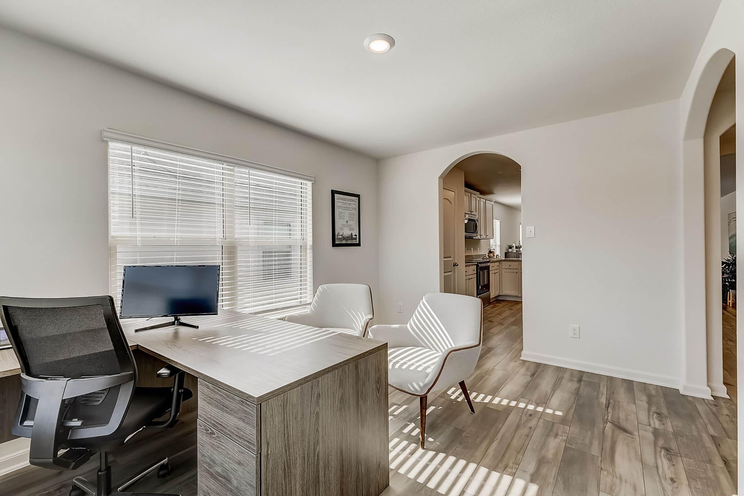 A modern home office featuring a sleek desk with a computer monitor, two white chairs, and large windows letting in natural light. The space has light-colored walls and wood flooring, creating a bright and airy atmosphere. In the background, a doorway leads to a kitchen area.