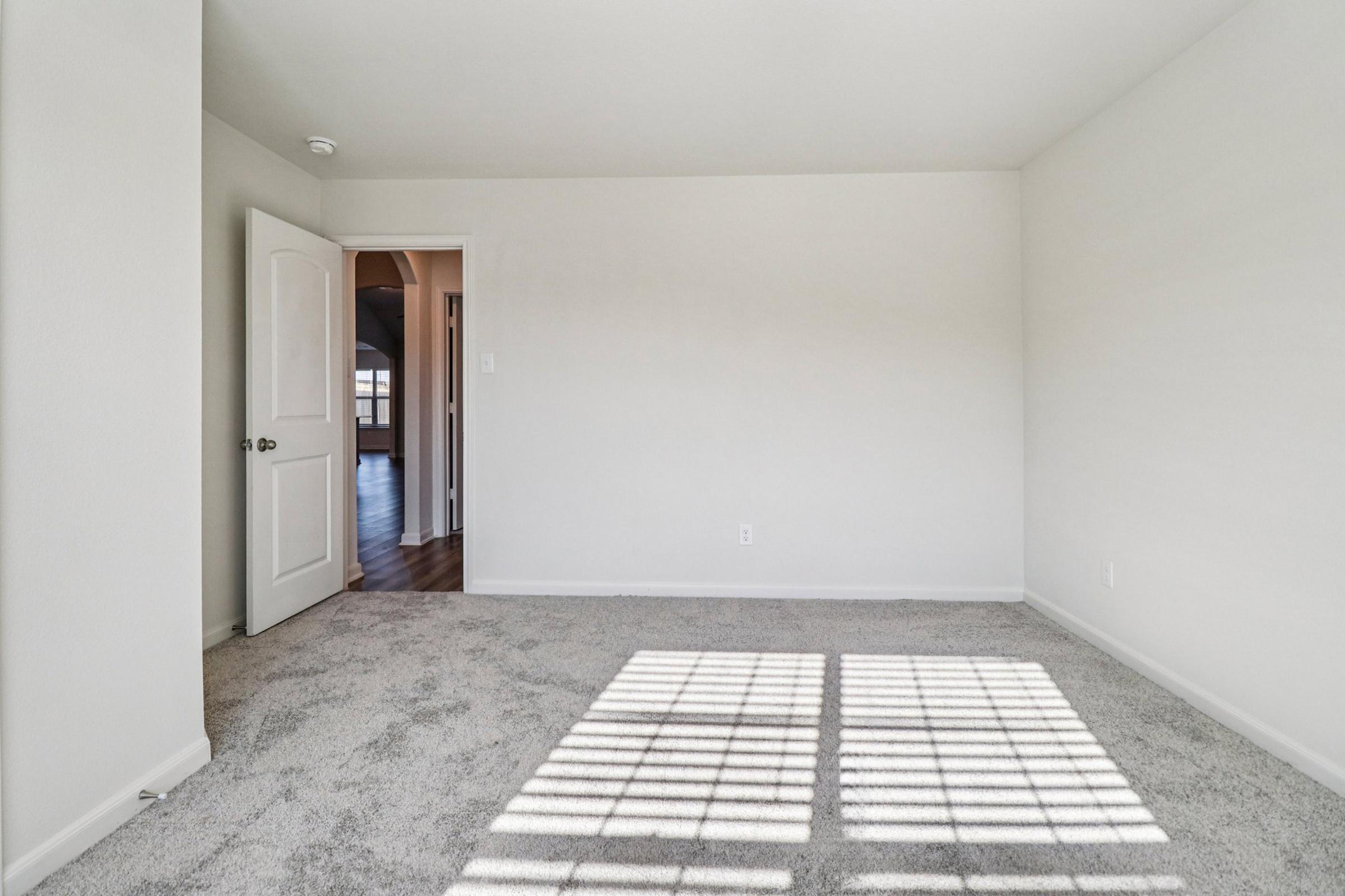 A spacious, empty room with light-colored walls and a plush, carpeted floor. A doorway leads to another area, and sunlight casts a patterned shadow on the floor from a nearby window. The room has a minimalist design, suitable for various furnishings.