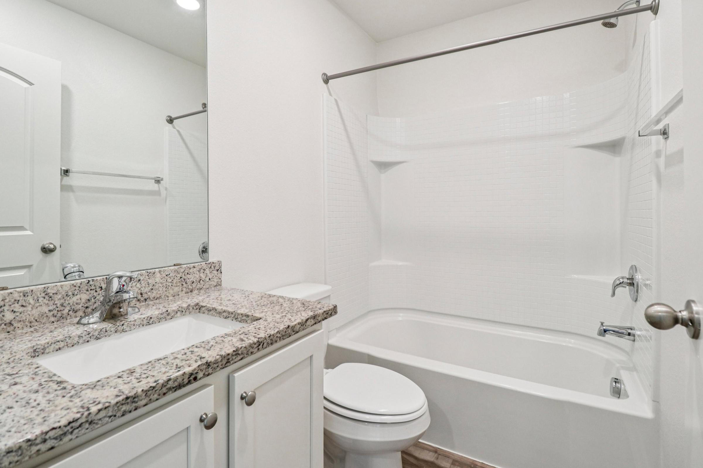 A clean and modern bathroom featuring a white tub and shower combo, a granite countertop sink with a chrome faucet, white cabinetry, and light-colored walls. The space is well-lit and minimalistic, enhancing its fresh appearance.