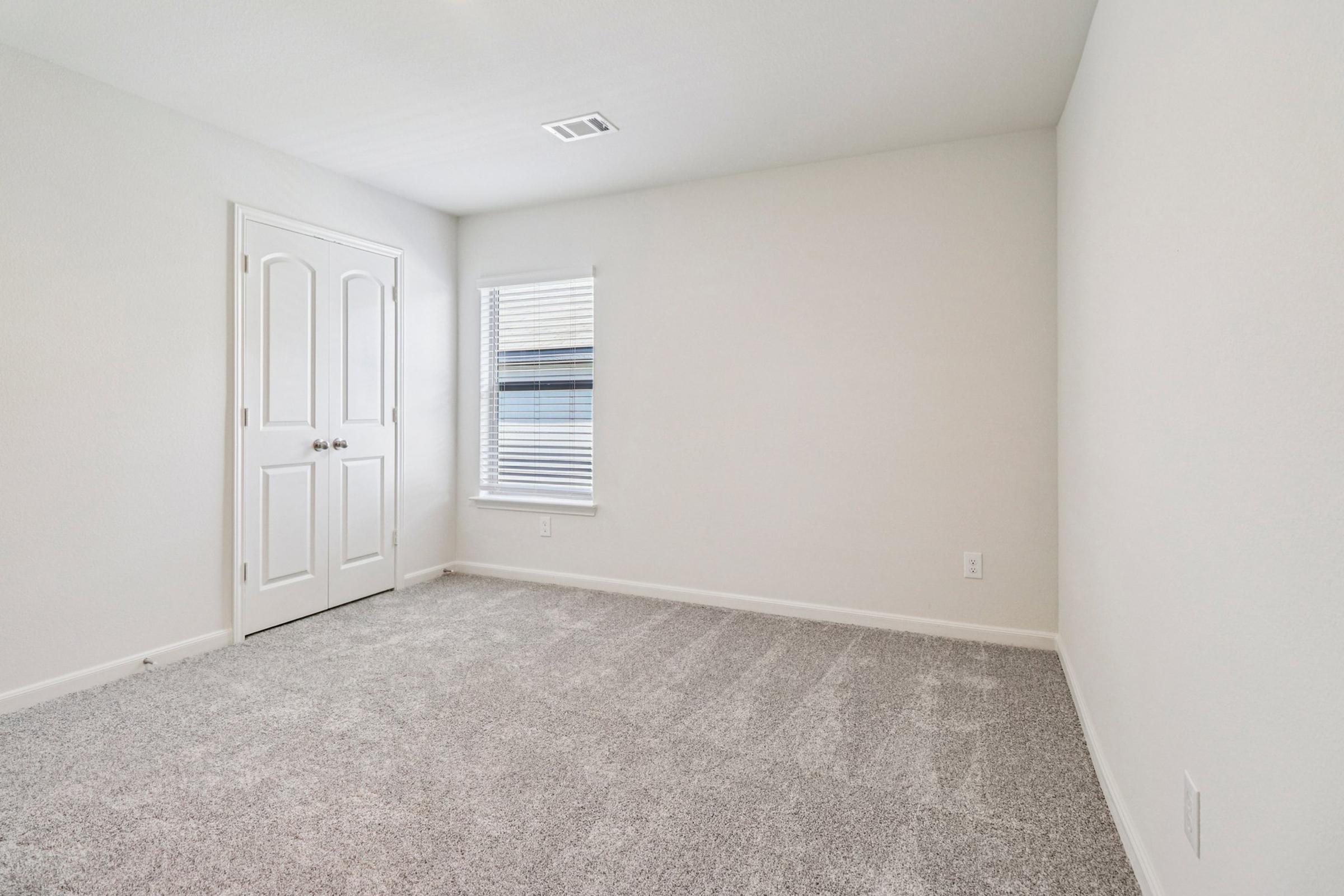 A vacant room with light grey carpet, a window with blinds, and a closed white door featuring two handles. The walls are painted a soft white, and the space is empty with no furniture, creating a bright and spacious feel. Natural light is entering through the window.