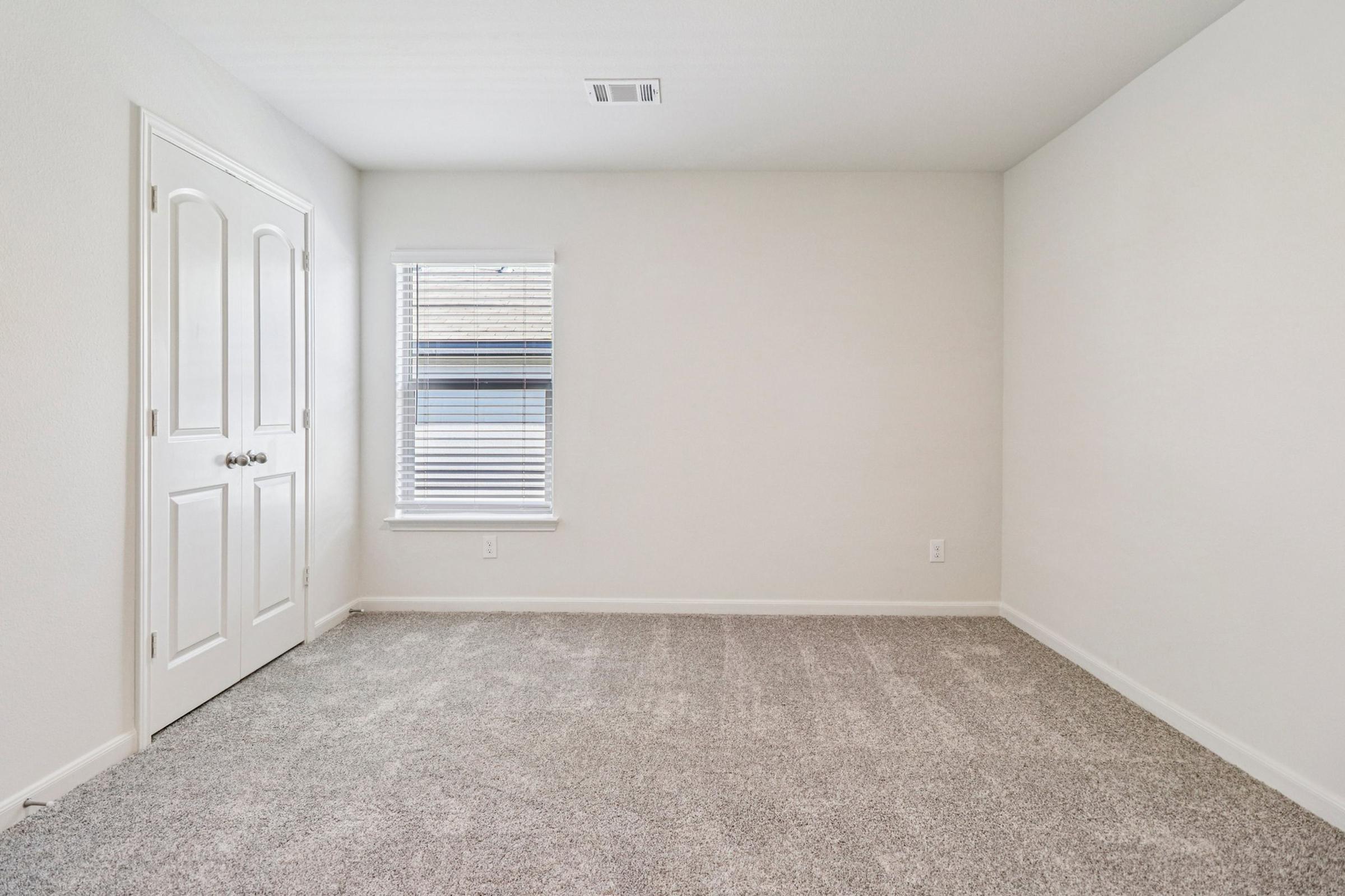 A clean, empty room with light gray carpet, white walls, and a single window with blinds. There is a closed door on the left with double handles, and no furniture is present, creating a spacious and minimalistic atmosphere.
