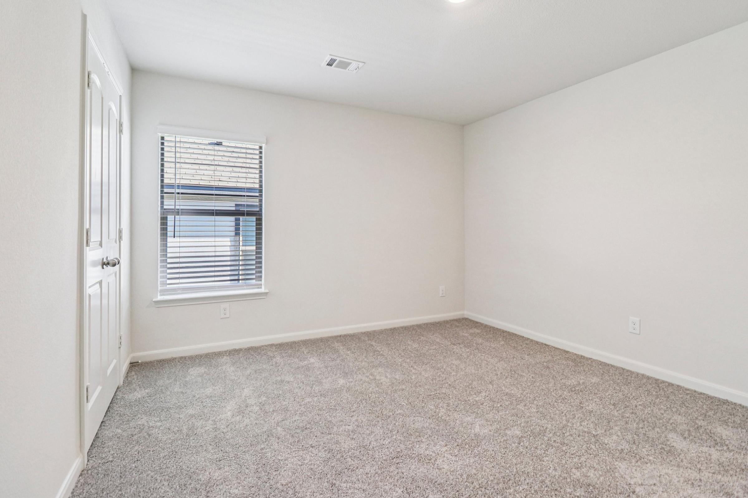 A vacant, well-lit room with light gray carpet and white walls. A window with horizontal blinds is visible on one wall, and a closed white door is on the opposite wall. The room has minimal furnishings, creating a clean, spacious feel.