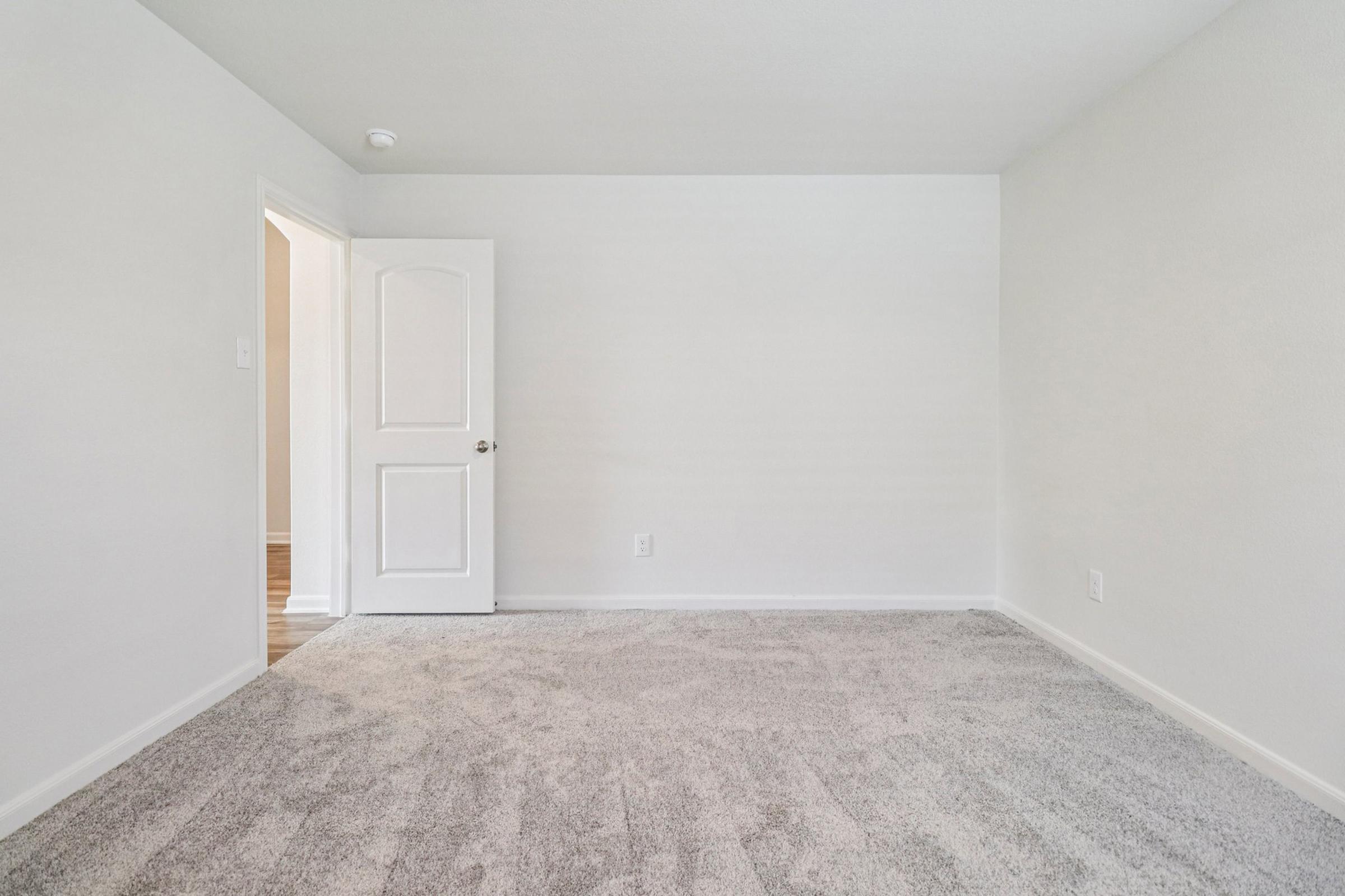 Empty room with light gray carpet and white walls. A closed white door on the left leads to another room, while the entrance to the hallway is visible in the background. The space is well-lit and uncluttered, creating a bright and open atmosphere.