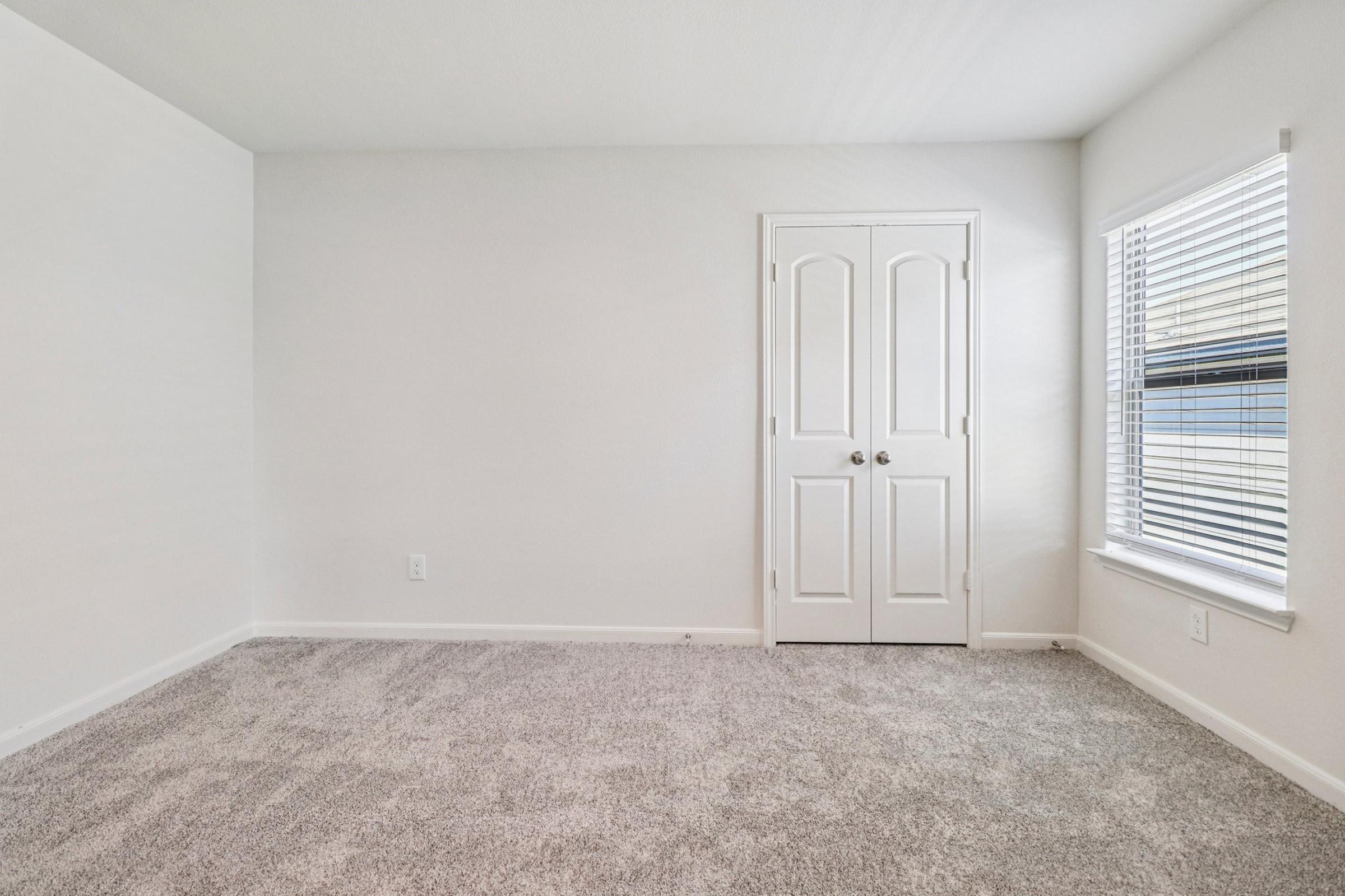 A clean, empty room featuring light gray carpeting, white walls, and a closed double-door closet. A window on the right side has white blinds, allowing natural light to enter the space, creating a bright and airy atmosphere. The overall appearance is minimalist and ready for personalization.