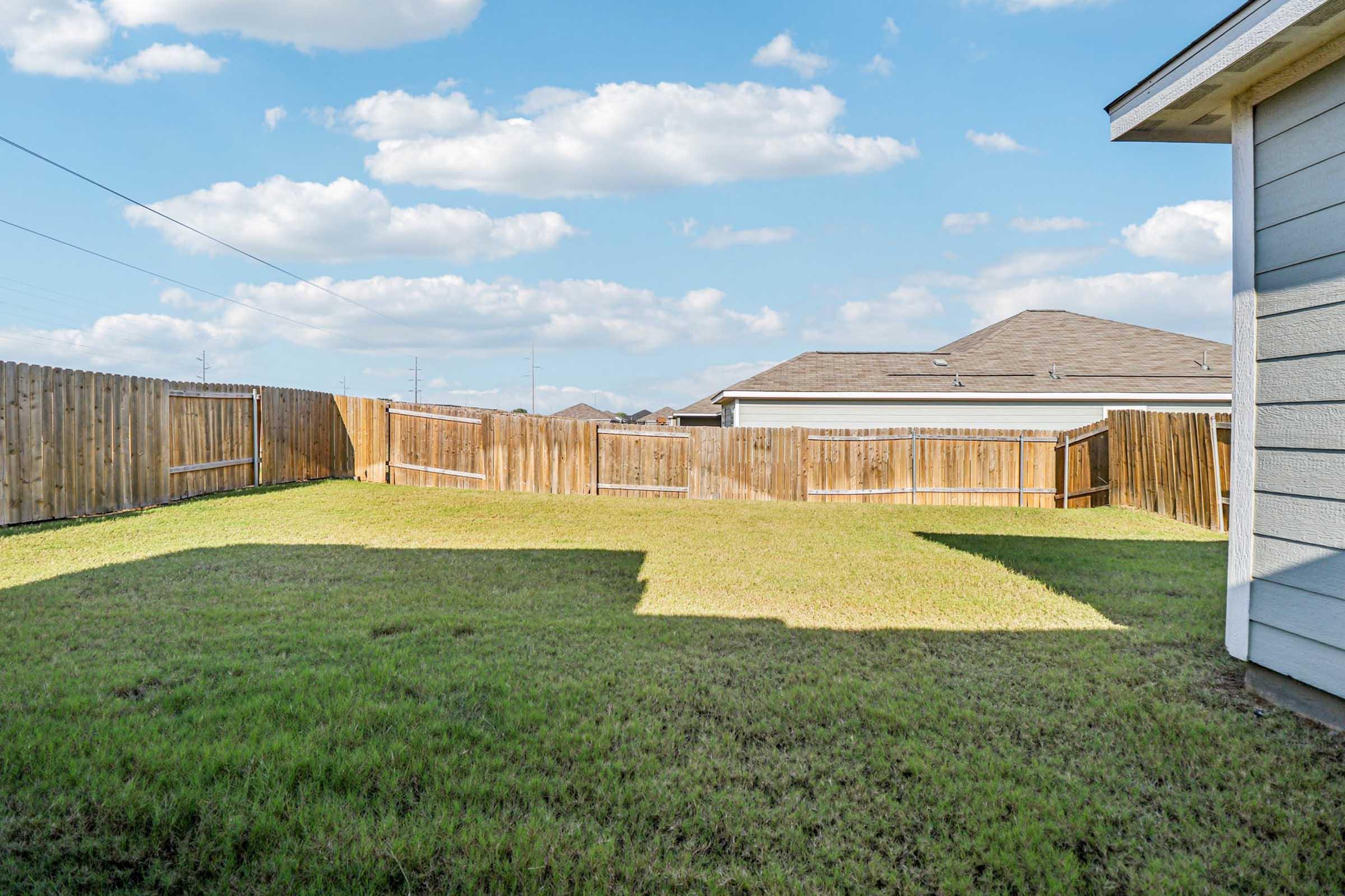 A clear, sunny backyard featuring a spacious grassy area bordered by wooden fences. In the background, residential homes are visible under a bright blue sky with scattered clouds. The image captures a peaceful outdoor space with a well-maintained lawn.