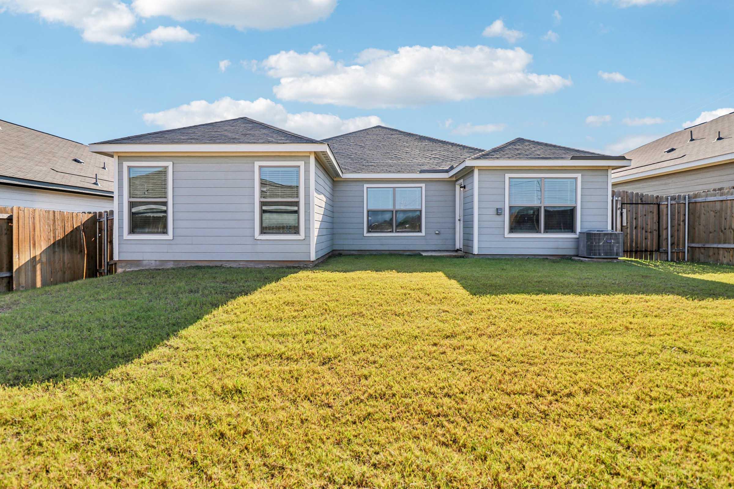 A spacious backyard featuring a well-maintained lawn and a single-story house with large windows. The house has a light-colored exterior and is surrounded by a wooden fence, with clear blue skies and a few clouds in the background.