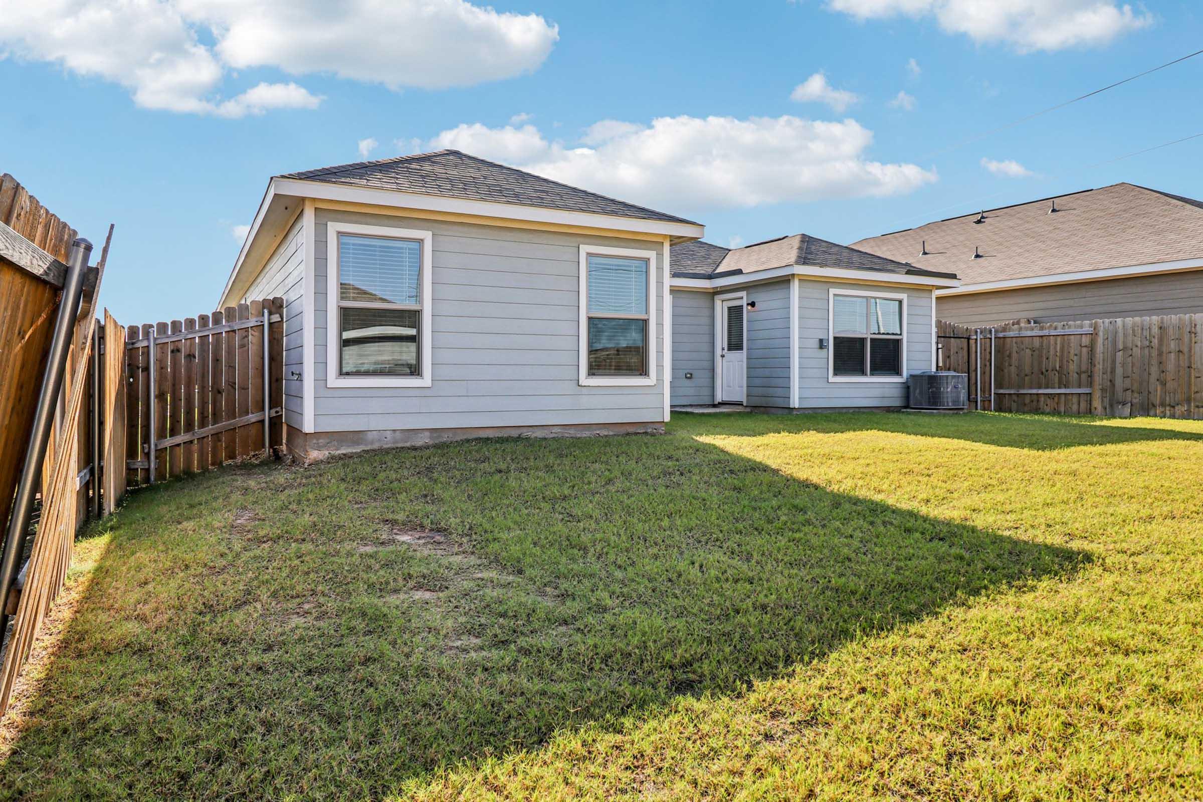 A view of a single-story house with light gray siding, featuring two front-facing windows. The backyard is spacious with lush green grass, surrounded by a wooden fence. A small air conditioning unit is visible beside the house, and a clear sky with a few clouds completes the scene.