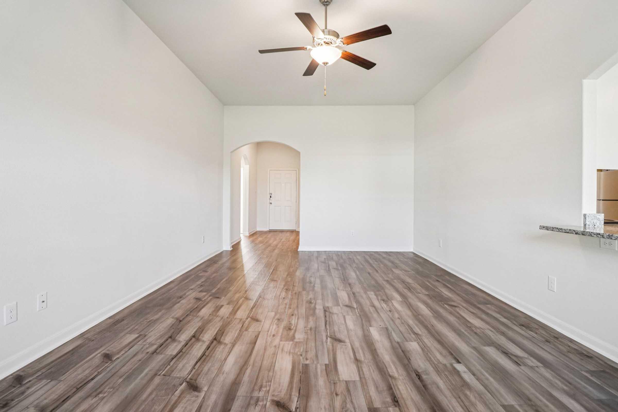 A spacious, well-lit interior with high ceilings and a ceiling fan. The room features a light-colored, smooth wall and a wooden floor. An arched doorway leads to a hallway, while a small counter area is visible on the right. Natural light fills the space, creating an inviting atmosphere.