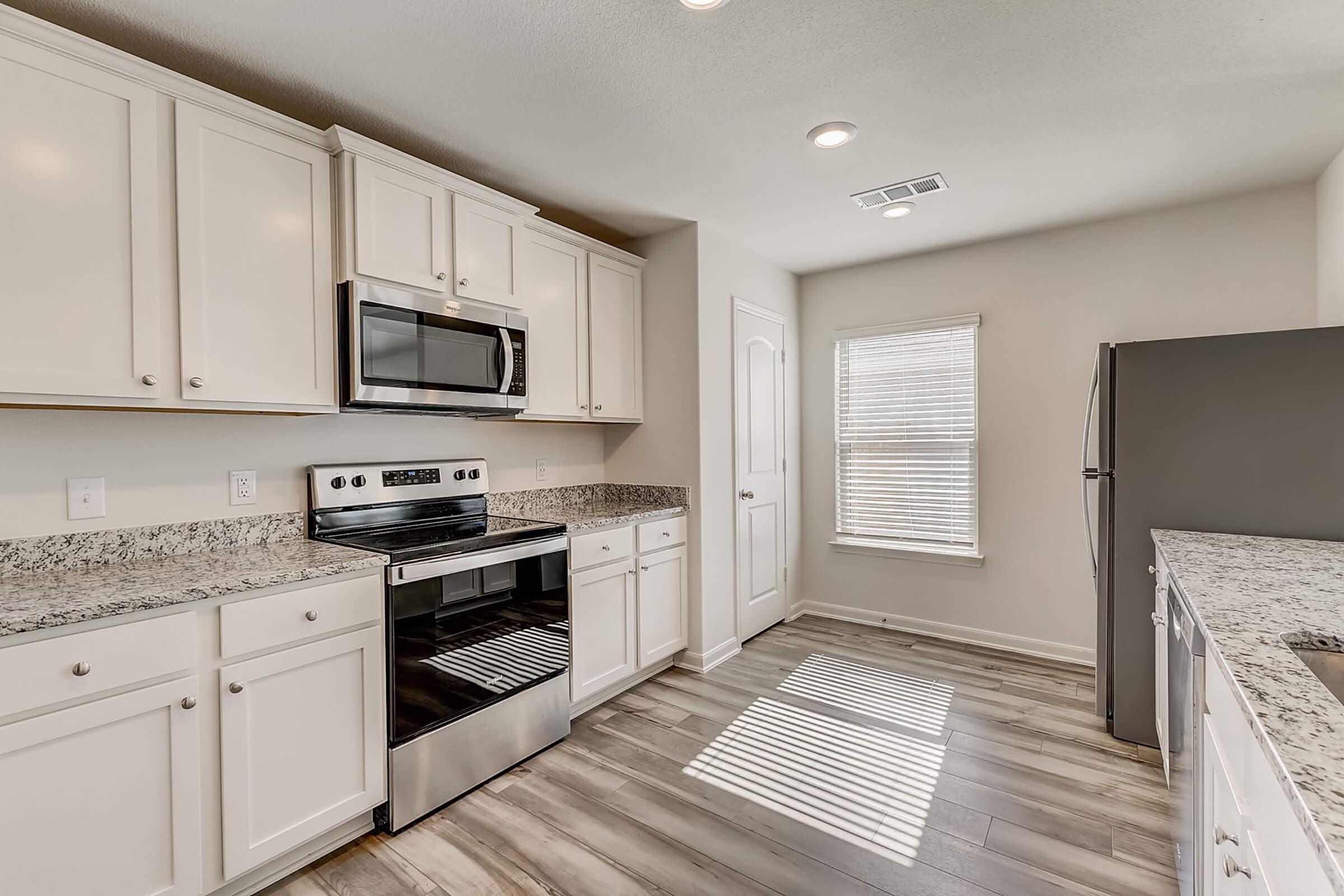 Modern kitchen with white cabinets, granite countertops, stainless steel appliances including an oven, microwave, and refrigerator. Natural light streams in through a window, casting shadows on the wooden floor. The space is clean and contemporary, perfect for cooking and dining.