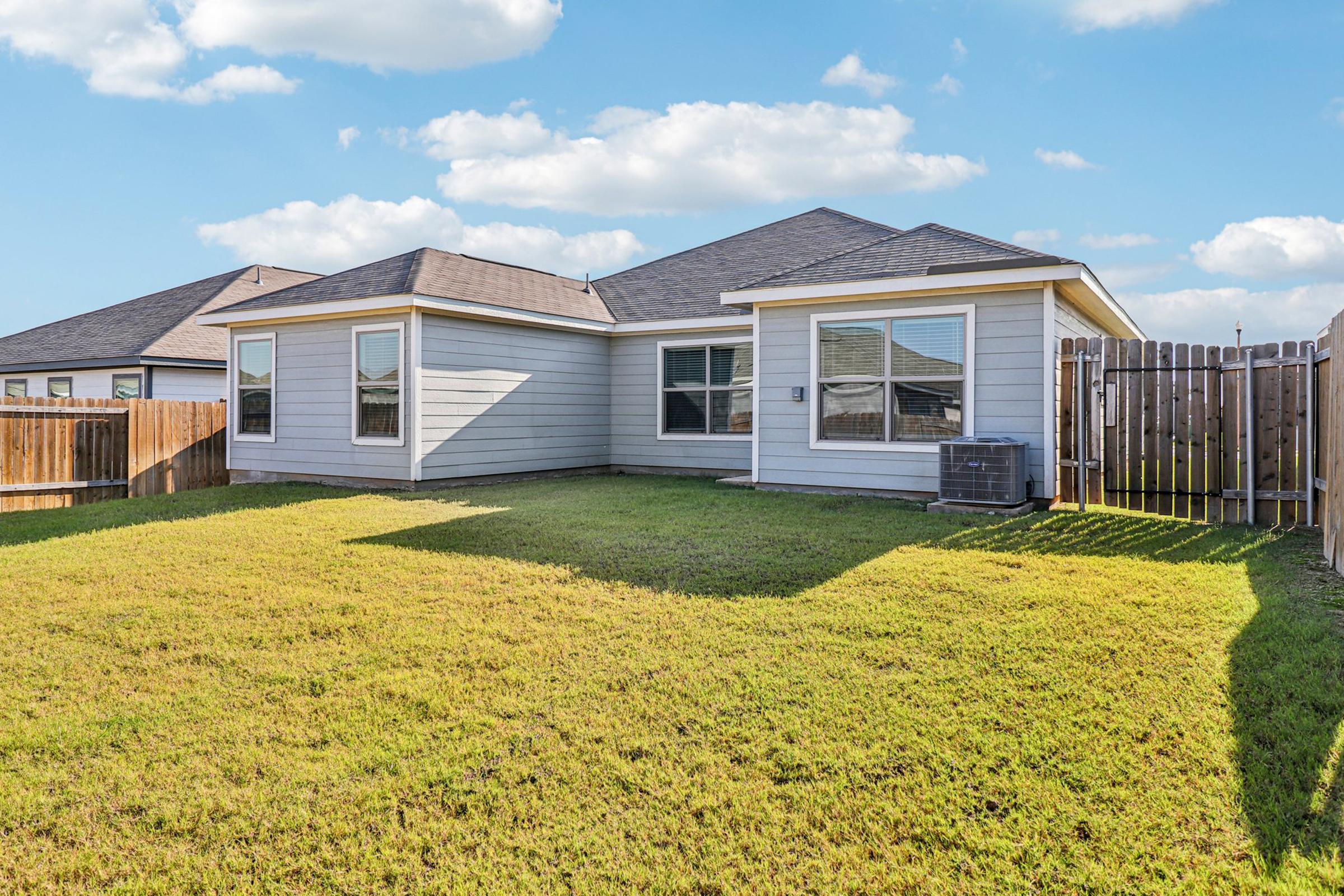 A gray house with a sloped roof sits on a green lawn, featuring multiple windows on one side. A wooden fence borders the yard, and an air conditioning unit is positioned next to the house. The sky is bright with fluffy clouds, creating a sunny atmosphere.