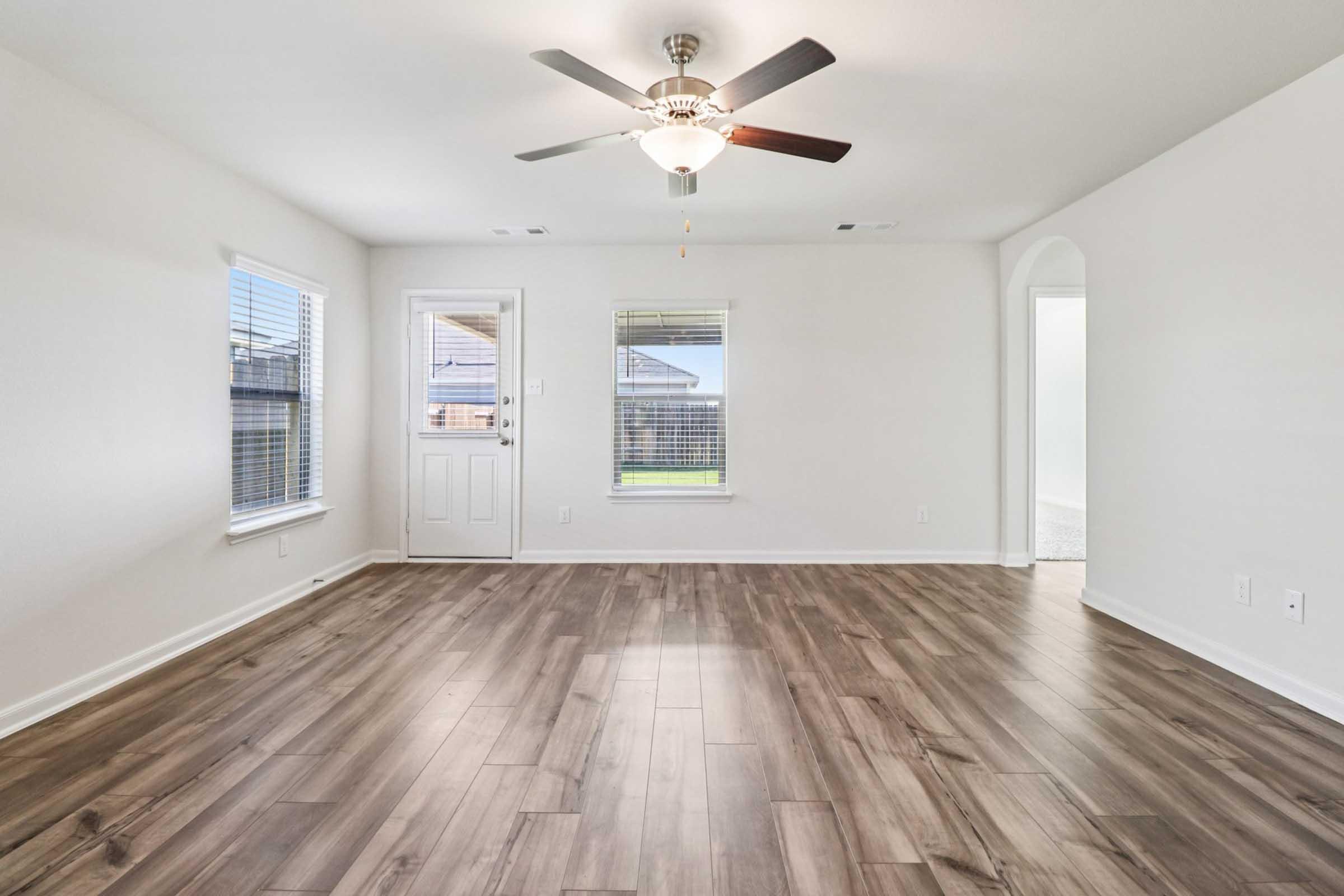 A spacious, well-lit living room featuring wood-look tile flooring, a ceiling fan, and large windows. One corner displays a front door leading outside. The room is painted in neutral colors, creating a bright and inviting atmosphere. An archway on the right leads to another space.