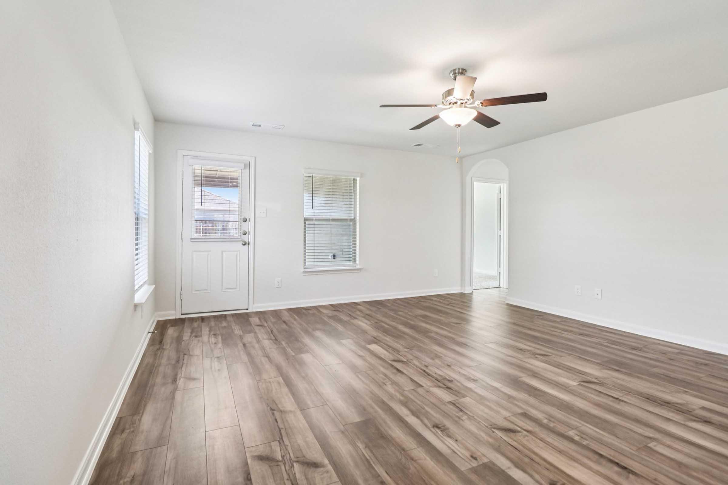 A spacious, well-lit living room featuring light-colored walls and a ceiling fan. The floor is covered with light brown wood-like tiles. There are two windows allowing natural light, and a door leading outside. The room has an open feel, with a hallway visible at the far end leading to other rooms.
