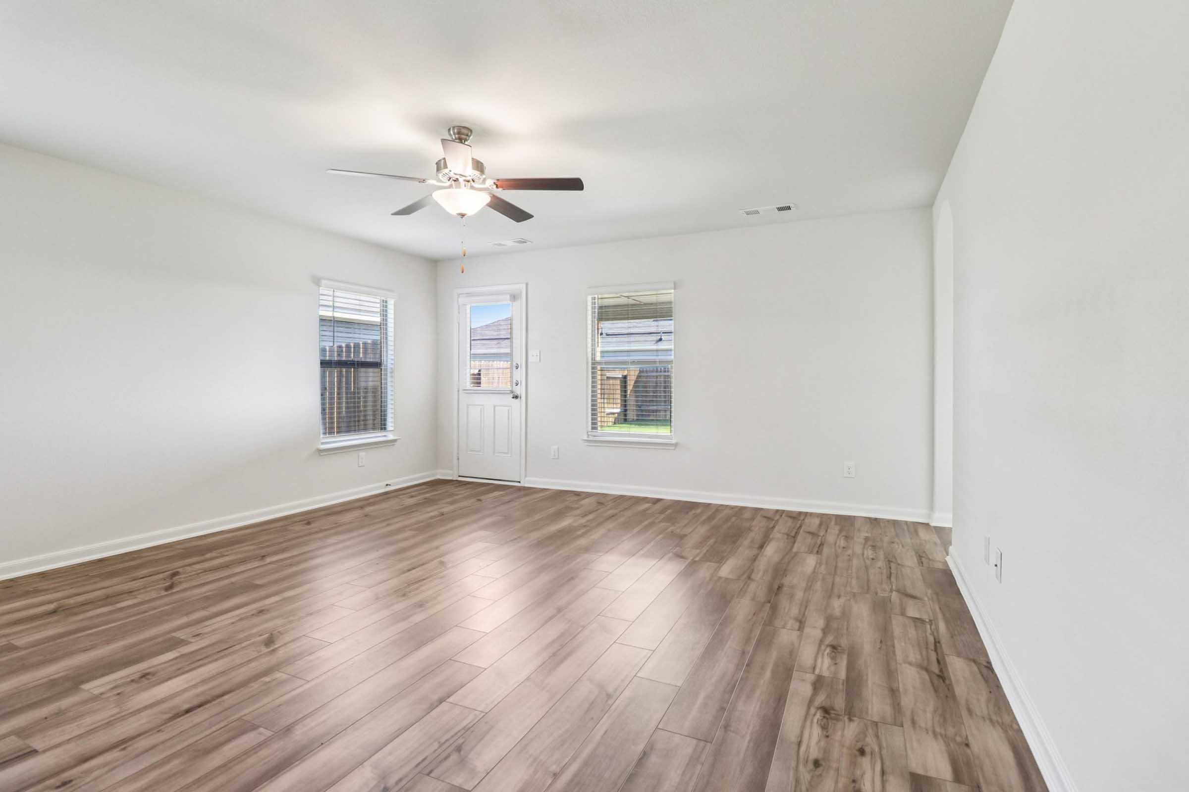 A well-lit, empty living room featuring light-colored walls, a ceiling fan, and wooden flooring. Two windows allow natural light to fill the space, while a front door is visible, leading outside. The minimalist design creates an open and airy atmosphere.