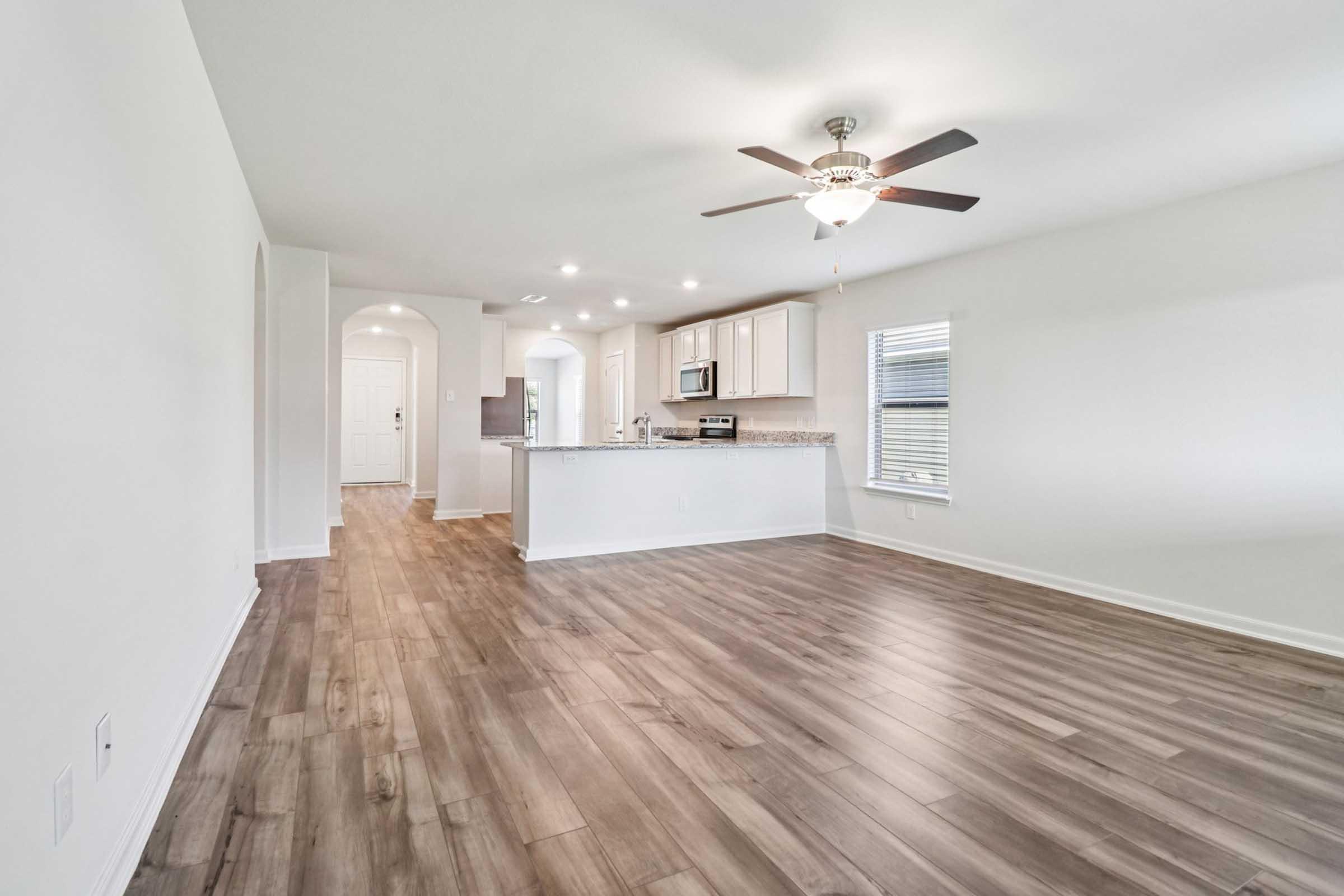 A spacious, modern living room featuring light-colored walls and large windows with natural light. The floor is made of laminated wood, and a ceiling fan hangs above. A kitchen area with white cabinets and countertops is visible in the background, along with an entryway leading to other areas of the home.