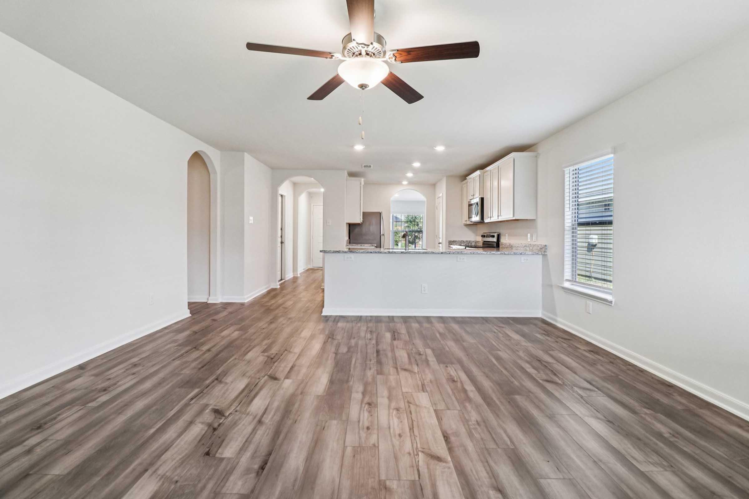 Spacious interior of a modern living area featuring light-colored walls, a ceiling fan, and large windows. The open layout connects to a kitchen with white cabinetry and a granite countertop, while hardwood-style flooring adds warmth and charm. Natural light fills the space, creating a welcoming atmosphere.