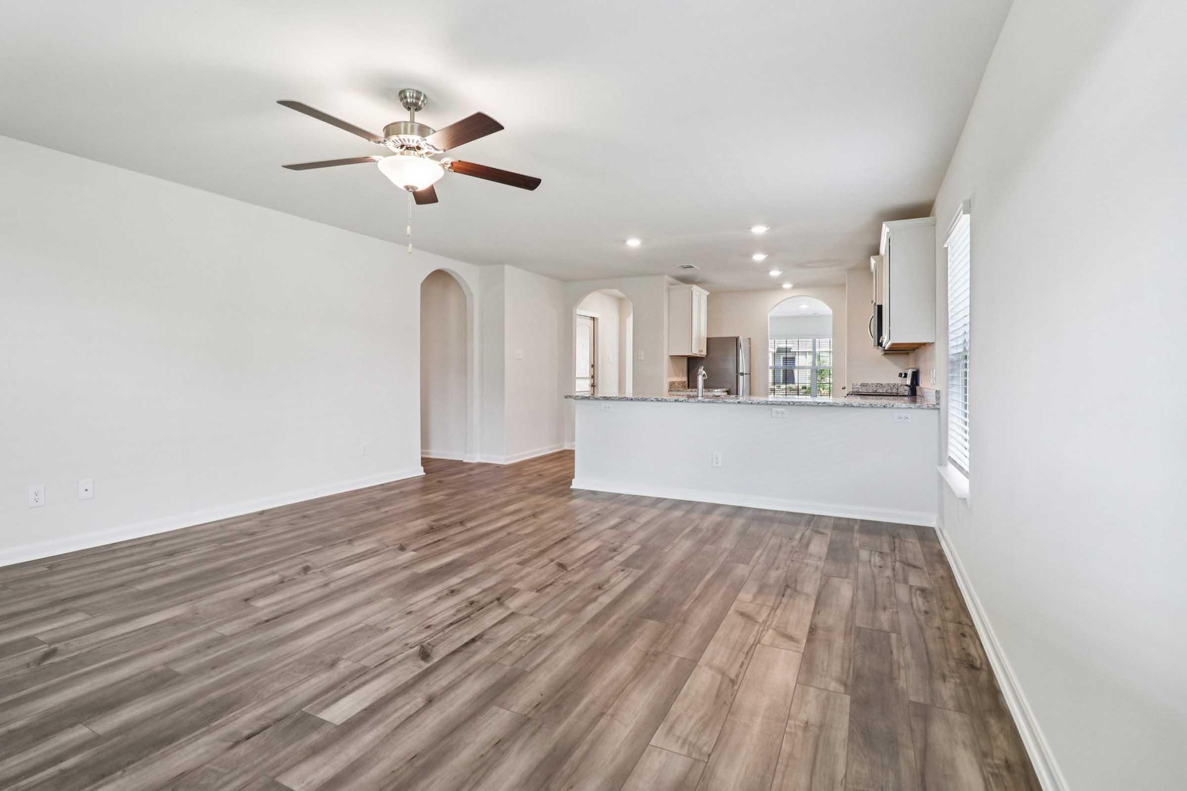A spacious, well-lit living area with light-colored walls and modern wood-style flooring. A ceiling fan is installed, and there's an open view into a kitchen featuring an archway. Natural light streams in through a window, enhancing the airy atmosphere.