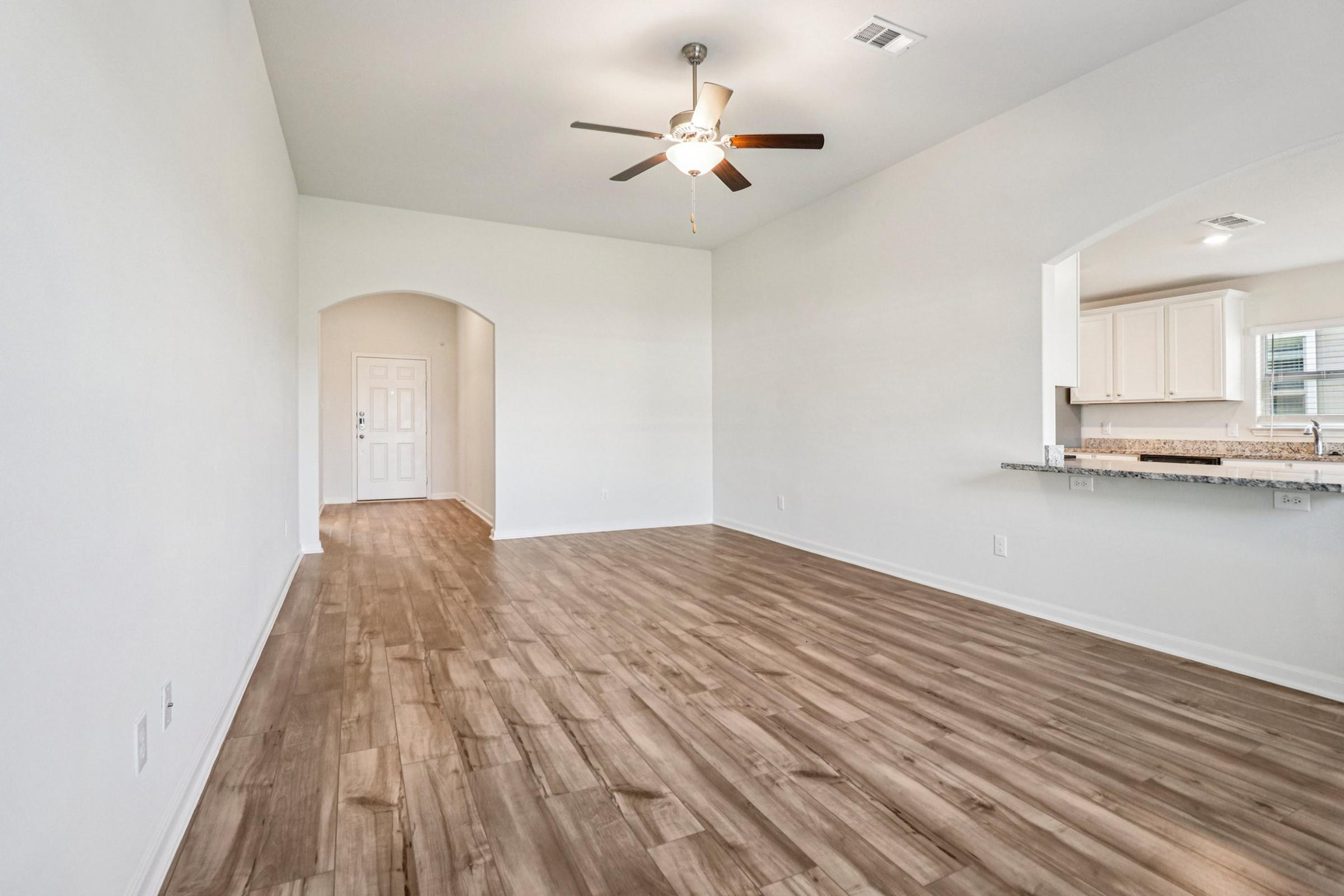 A spacious, light-filled living area with wooden flooring, featuring a ceiling fan and an opening to a kitchen. The walls are painted white, and there is a doorway leading to the entrance. The space is uncluttered, emphasizing a modern and airy atmosphere.