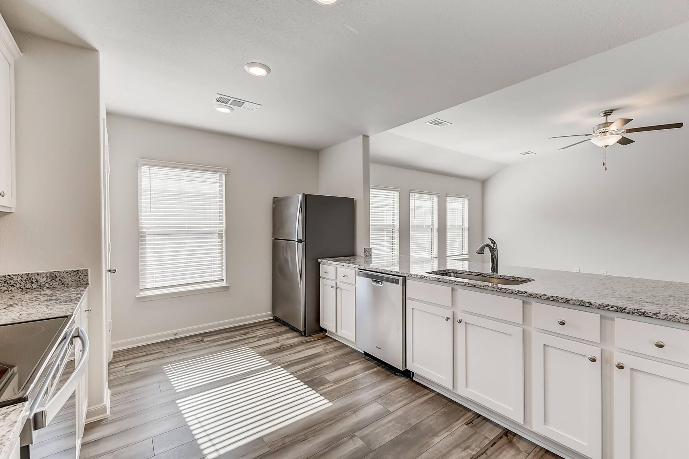 Bright, modern kitchen featuring stainless steel appliances, white cabinetry, and a granite countertop. Natural light streams in through multiple windows, highlighting the open layout and sleek design. The floor is adorned with wood-like tiles, creating a warm, inviting atmosphere.