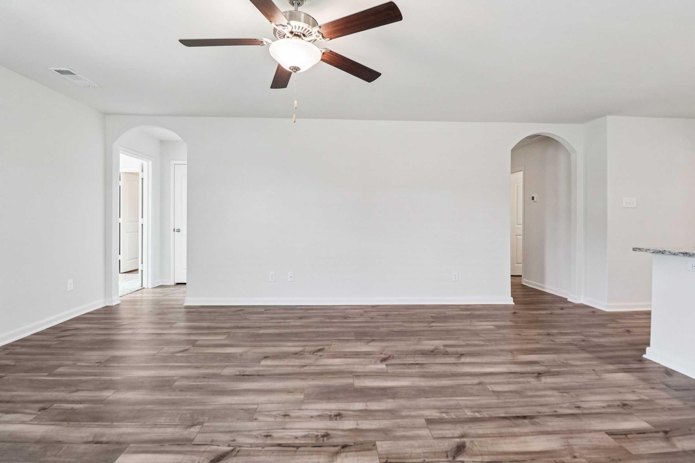 A spacious, unfurnished living room featuring light-colored walls and a dark wood floor. There is a ceiling fan with wooden blades, and arched doorways lead to adjacent rooms. Natural light floods the space, creating an inviting atmosphere.