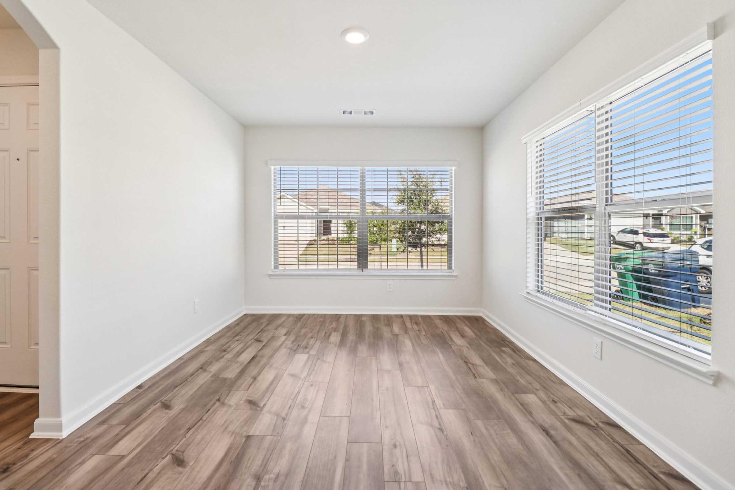 Empty room with light-colored walls and large windows featuring white blinds. The flooring is wood-like, with natural light streaming in. A view of greenery and parked cars can be seen outside through the windows. The room is spacious and uncluttered, creating a bright and inviting atmosphere.