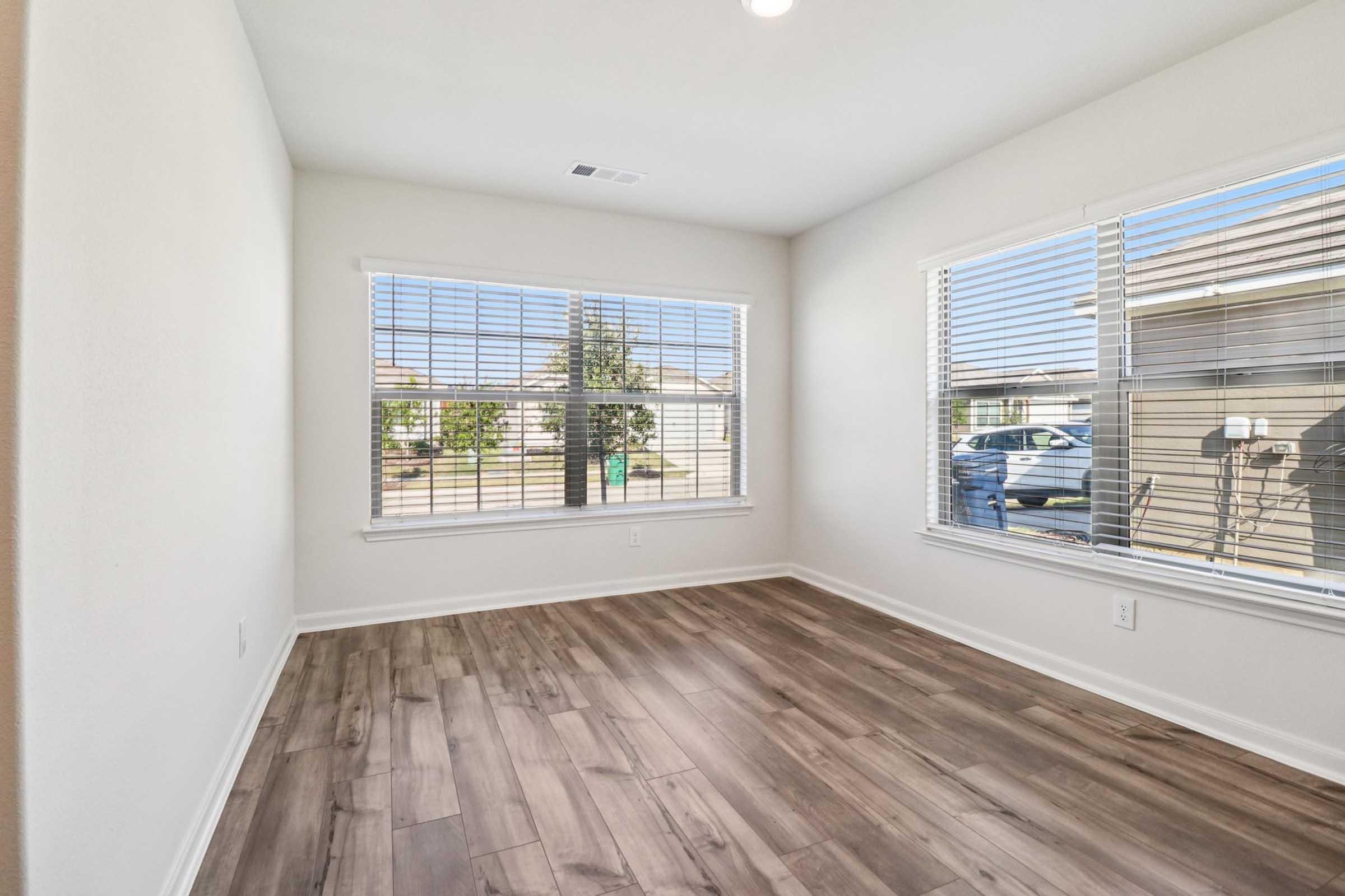 A bright, empty room with large windows on two walls, featuring white walls and wooden flooring. Sunlight streams in, illuminating the space. Outside the windows, there are glimpses of greenery and parked vehicles. The room is ready for furniture and decoration.