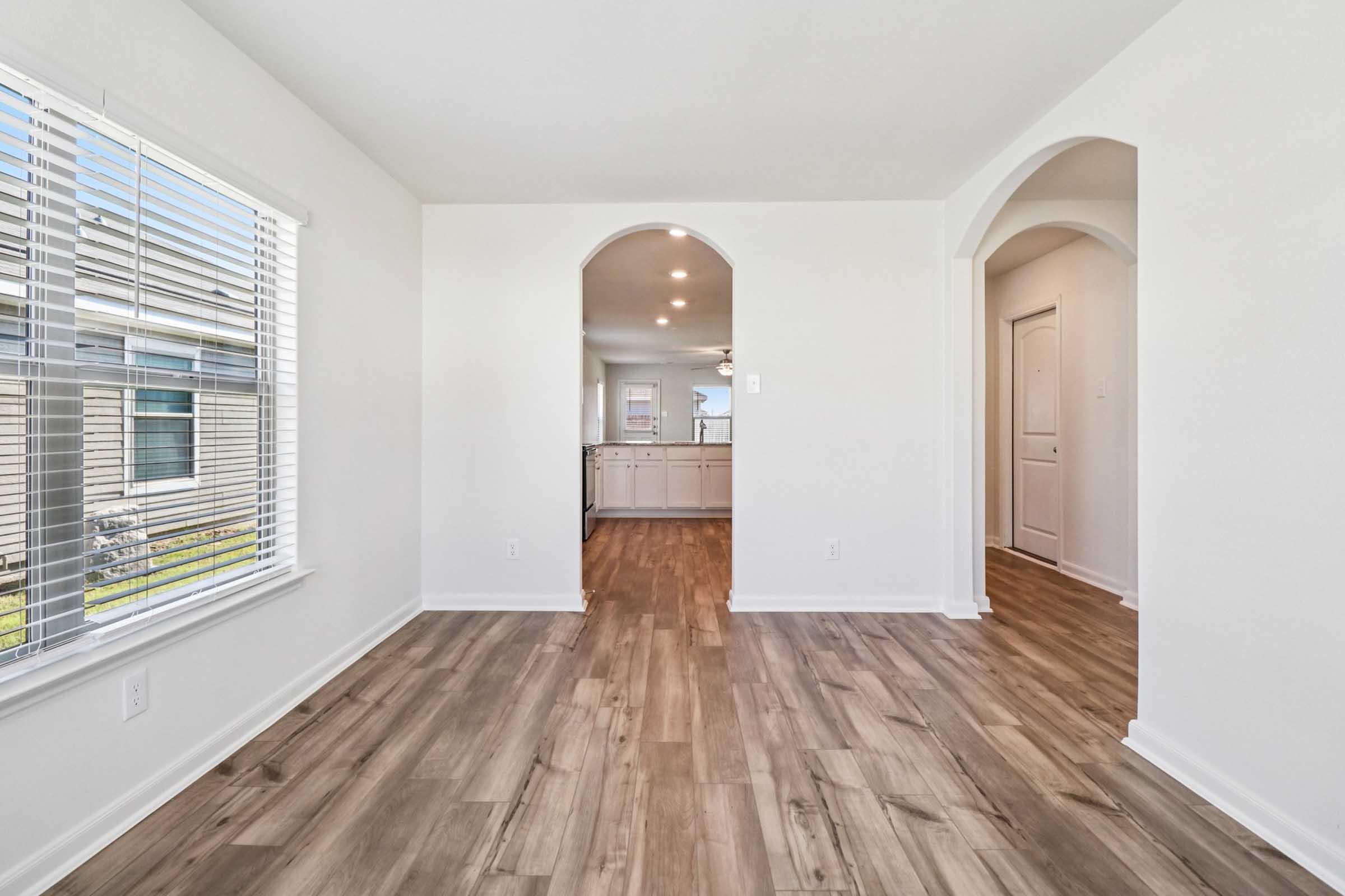 Interior of a modern, bright room featuring light wood flooring and white walls. The space includes large windows with blinds, two archways leading to adjoining rooms, and recessed ceiling lights. The layout is open and airy, creating a welcoming atmosphere.