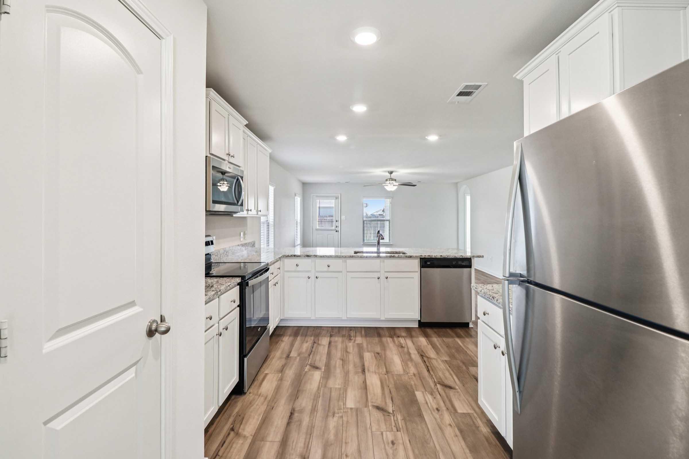 A modern kitchen featuring white cabinetry, stainless steel appliances, and granite countertops. The space includes a refrigerator, dishwasher, and stove, with a view towards a window. The flooring is a warm-toned wood, and there is ample natural light from the windows.