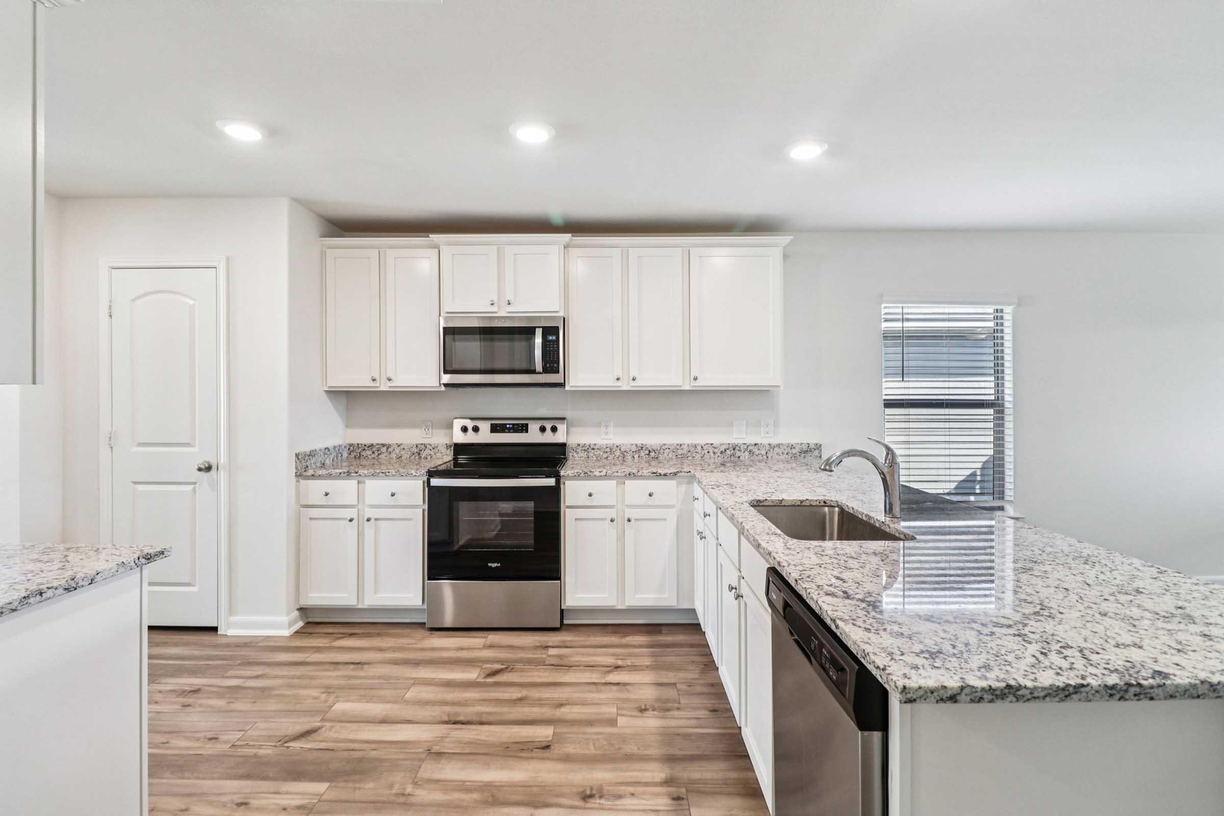 Modern kitchen featuring white cabinetry, granite countertops, and stainless steel appliances, including an oven, microwave, and dishwasher. The space has natural light from a window and light wood flooring, creating a bright and inviting atmosphere.