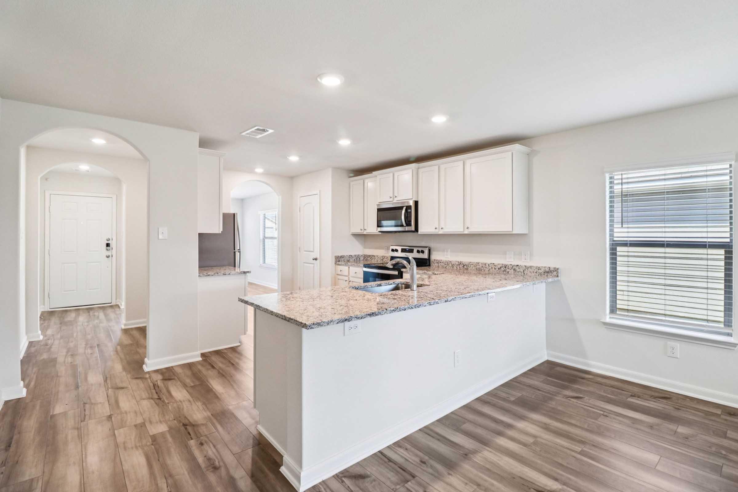 A modern kitchen with a large central island featuring a granite countertop, white cabinetry, stainless steel appliances, and a spacious layout. The room has an open floor plan, with windows allowing natural light, and wood-like flooring throughout.