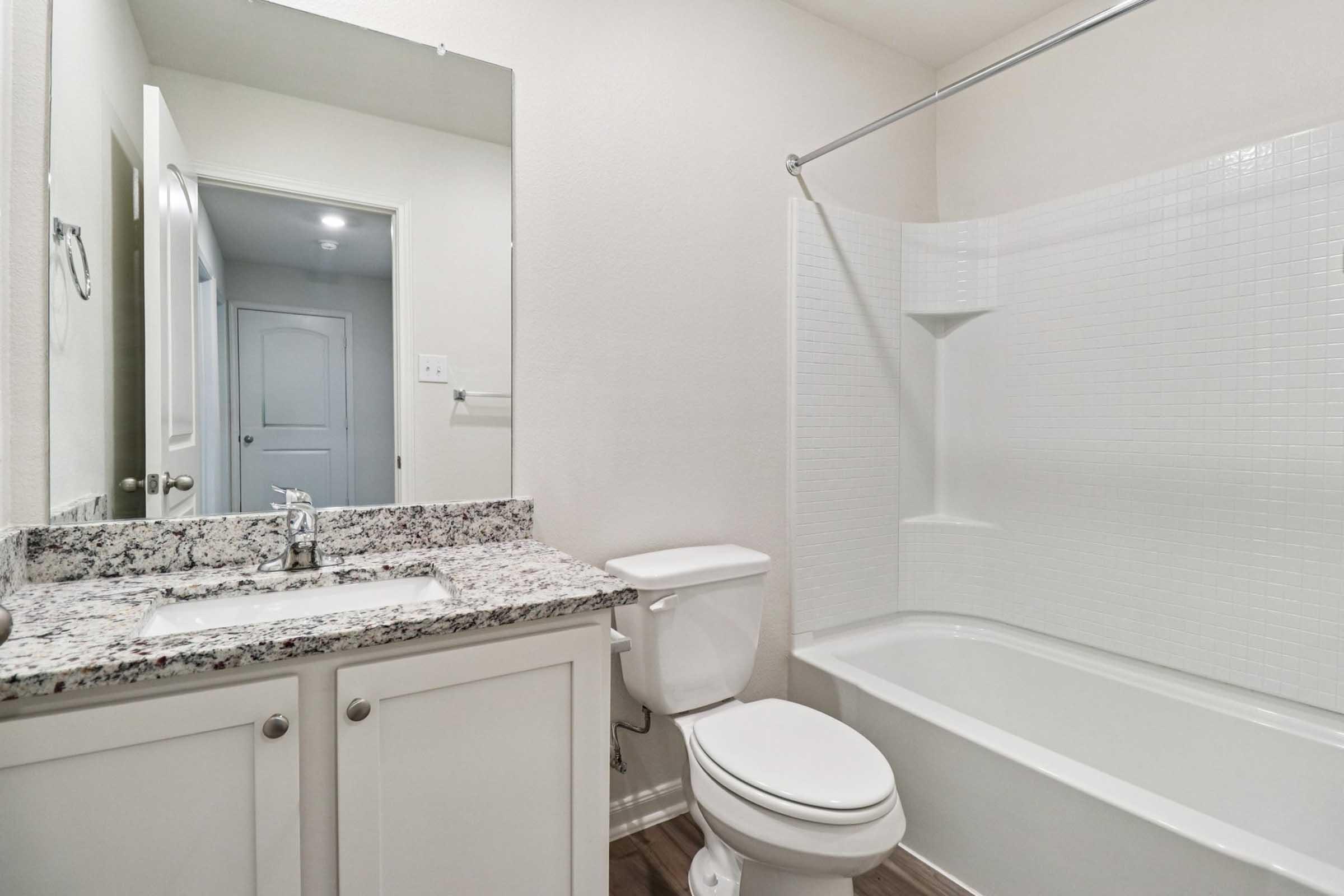 A clean and modern bathroom featuring a white bathtub with a shower curtain, a toilet, and a granite countertop vanity with a sink. The walls are painted in a light color, and there is a door leading to another room.