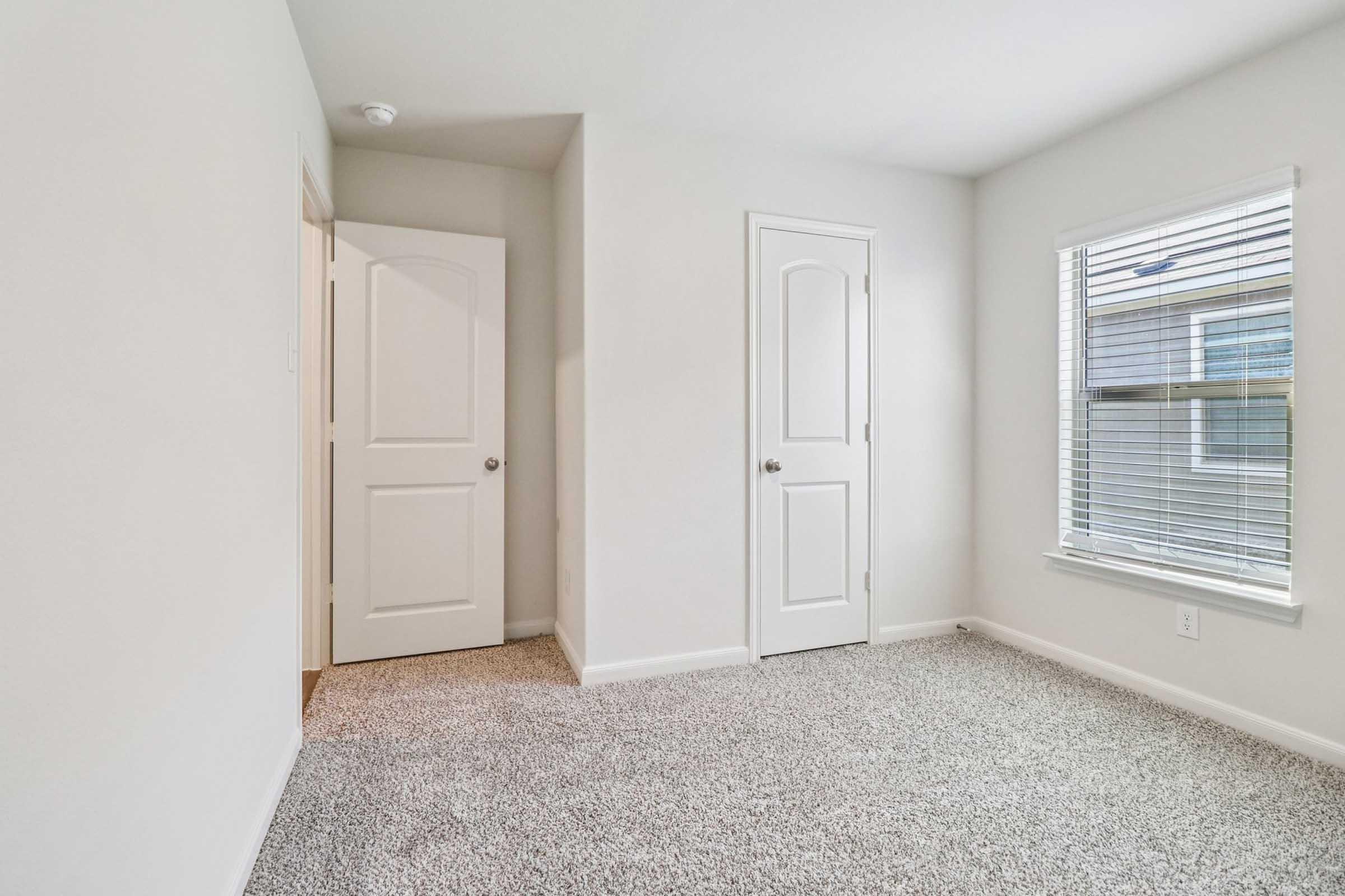 A small, empty room with light beige carpet and neutral-colored walls. There are two closed doors on the left side—one leading to a hallway and the other possibly a closet. A window on the right allows natural light to enter, providing a view of the outside.