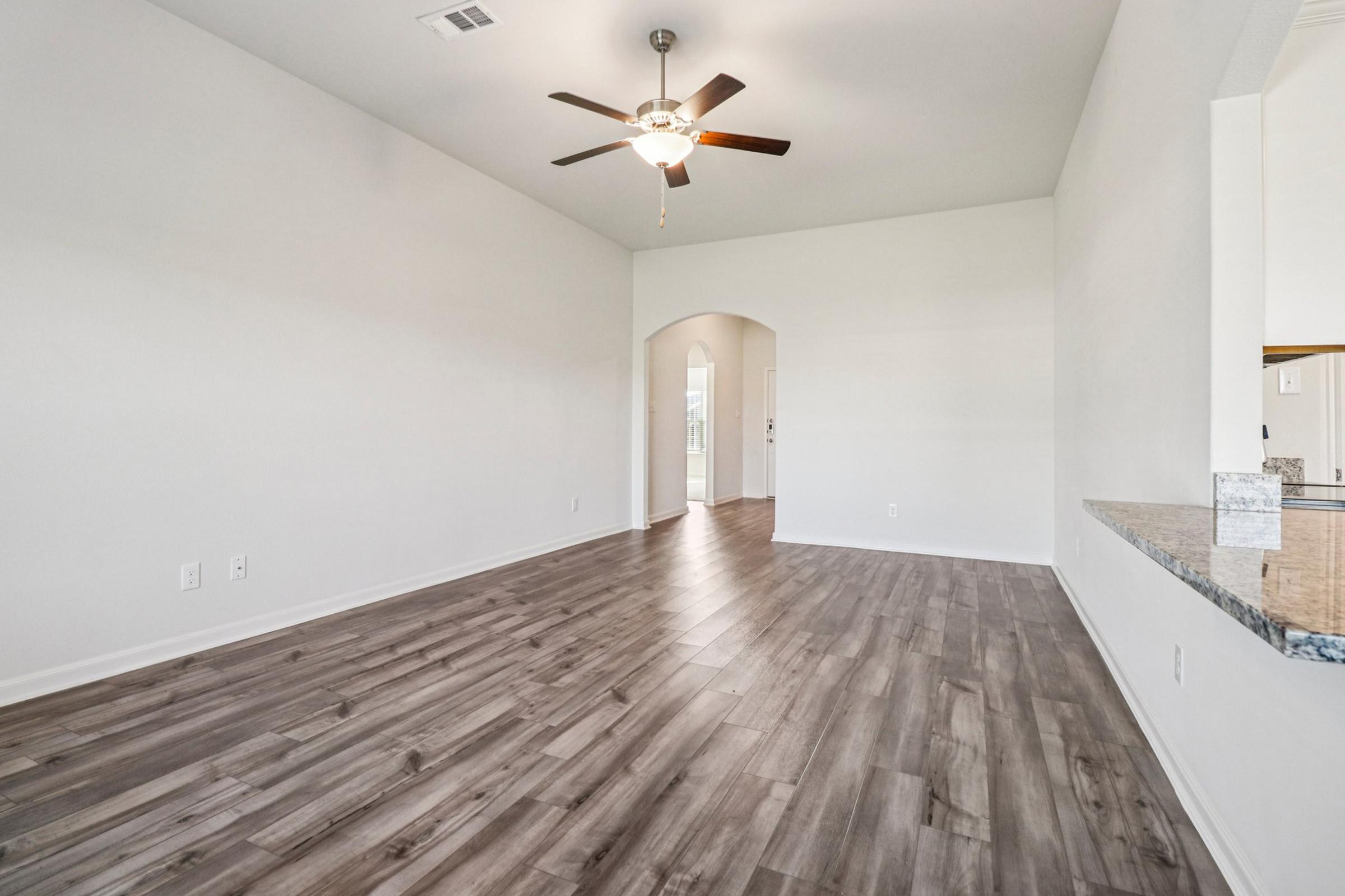 A spacious, empty living room with light gray walls and a ceiling fan. The flooring is a medium wood tone. An arched doorway leads to another room, and there is a counter on the right, likely a kitchen bar area. Natural light brightens the space through the doorway.
