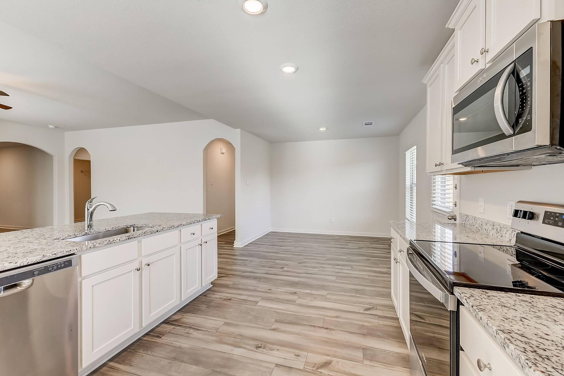 Modern kitchen featuring white cabinetry, granite countertops, stainless steel appliances, and wood-look flooring. The space has an open layout with natural light coming through windows and a ceiling fan. It includes a large island with sink and dishwasher, creating a functional cooking area.