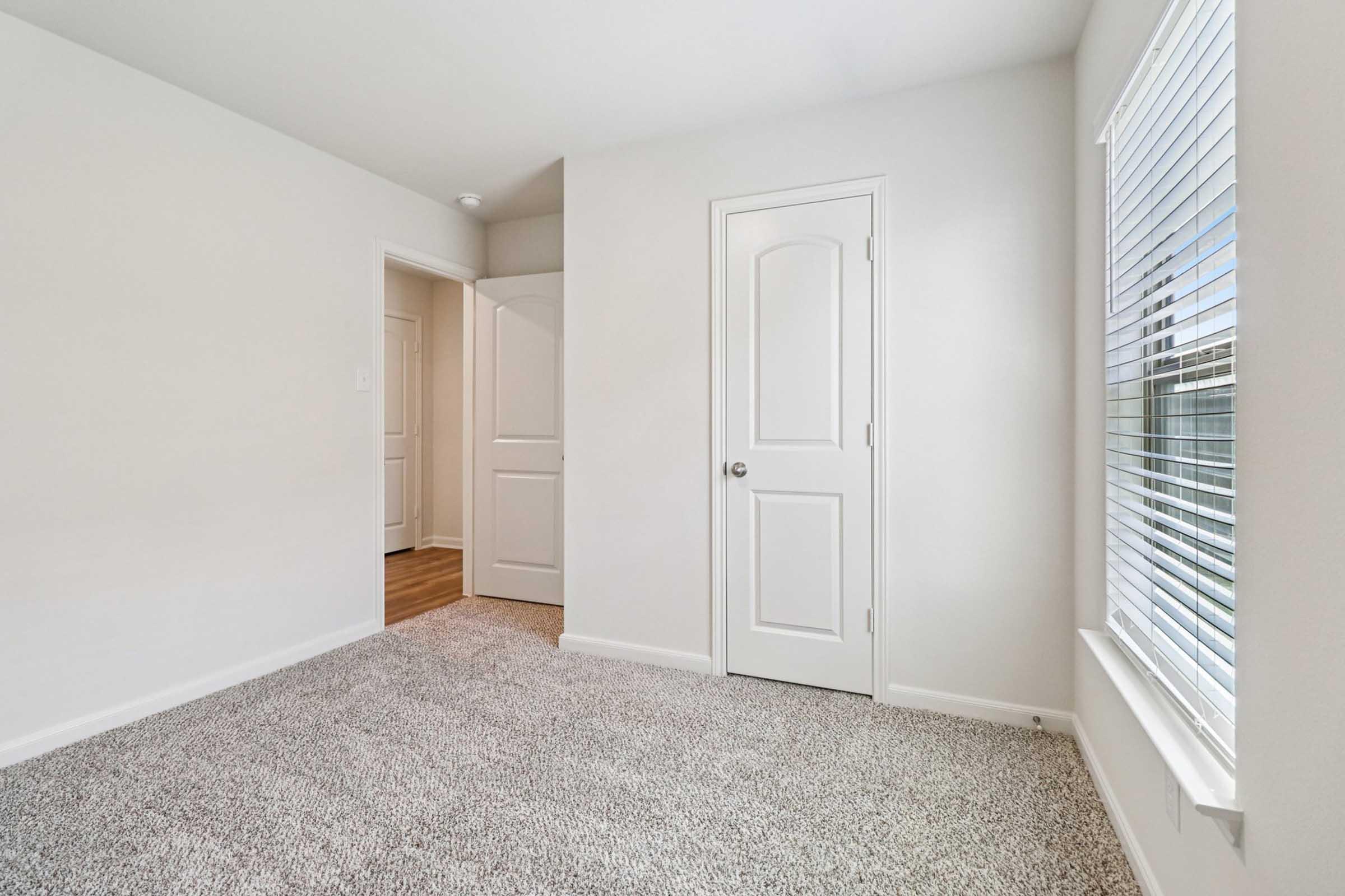 Empty room with light-colored walls and beige carpet. There is a closed door on the left and a window on the right with white blinds. The space is well-lit and appears neat and clean, suitable for various uses.