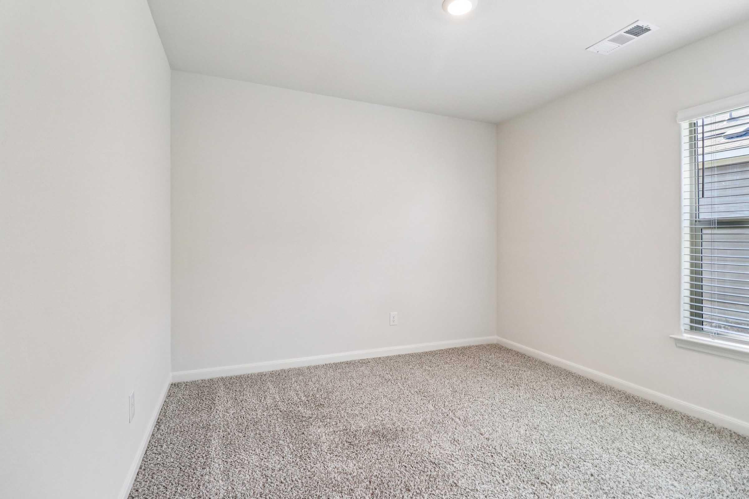 Empty room with light beige walls, carpeted floor in a similar tone, and a single window with blinds allowing natural light to enter. The space is simple and uncluttered, creating a neutral atmosphere.