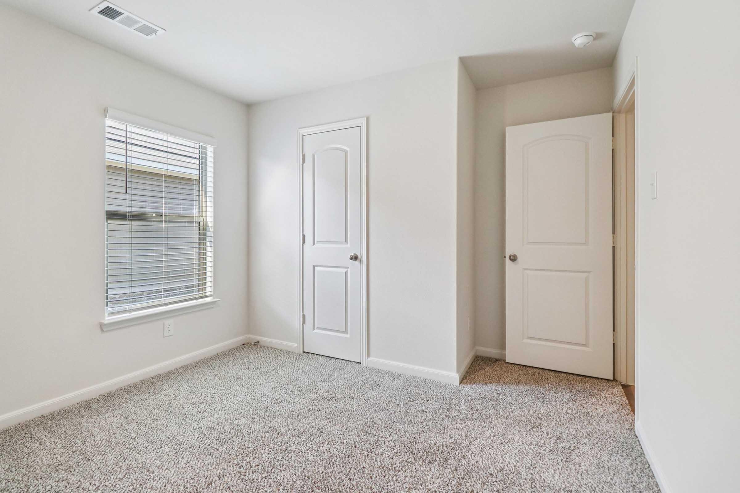 A bright and empty room with light gray walls and carpet. There is a window with blinds on the left, allowing natural light in. Two doors are visible, one closed and one slightly ajar, leading to another space. The room has a clean, minimalistic design with no furniture.