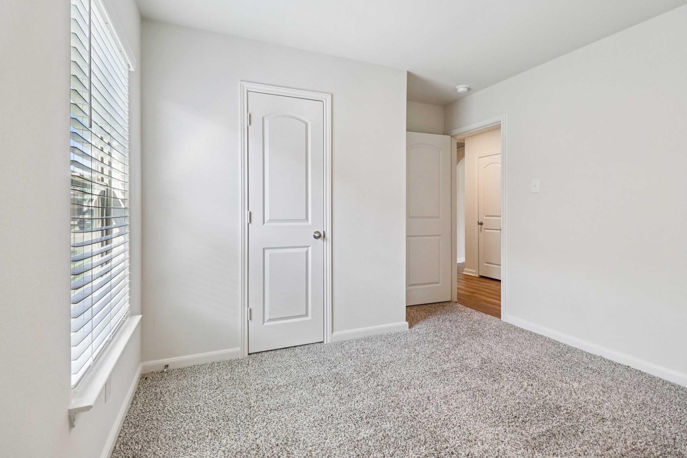Empty room with light gray walls and carpeted floor. A door on the left leads to another space, while a window with blinds allows natural light. The simple, neutral decor creates a bright and airy atmosphere.
