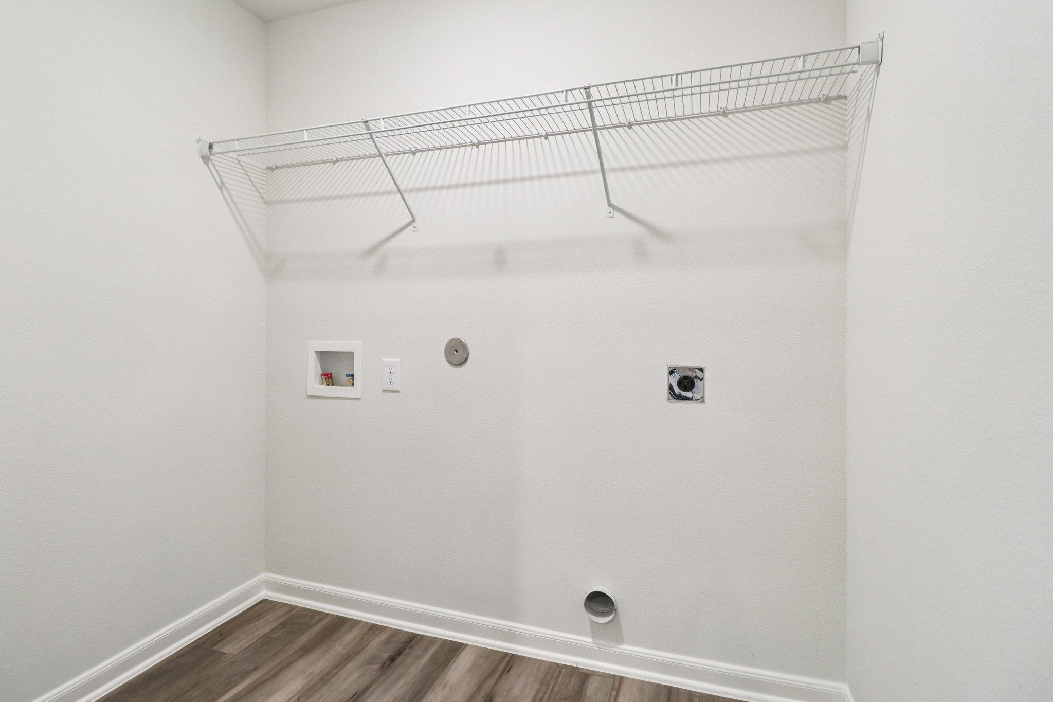 An empty laundry room featuring a white wire shelf mounted on a light-colored wall. There are small fixtures for electrical and plumbing connections on the wall. The floor is made of wood-like laminate, adding a warm touch to the otherwise minimalist space.