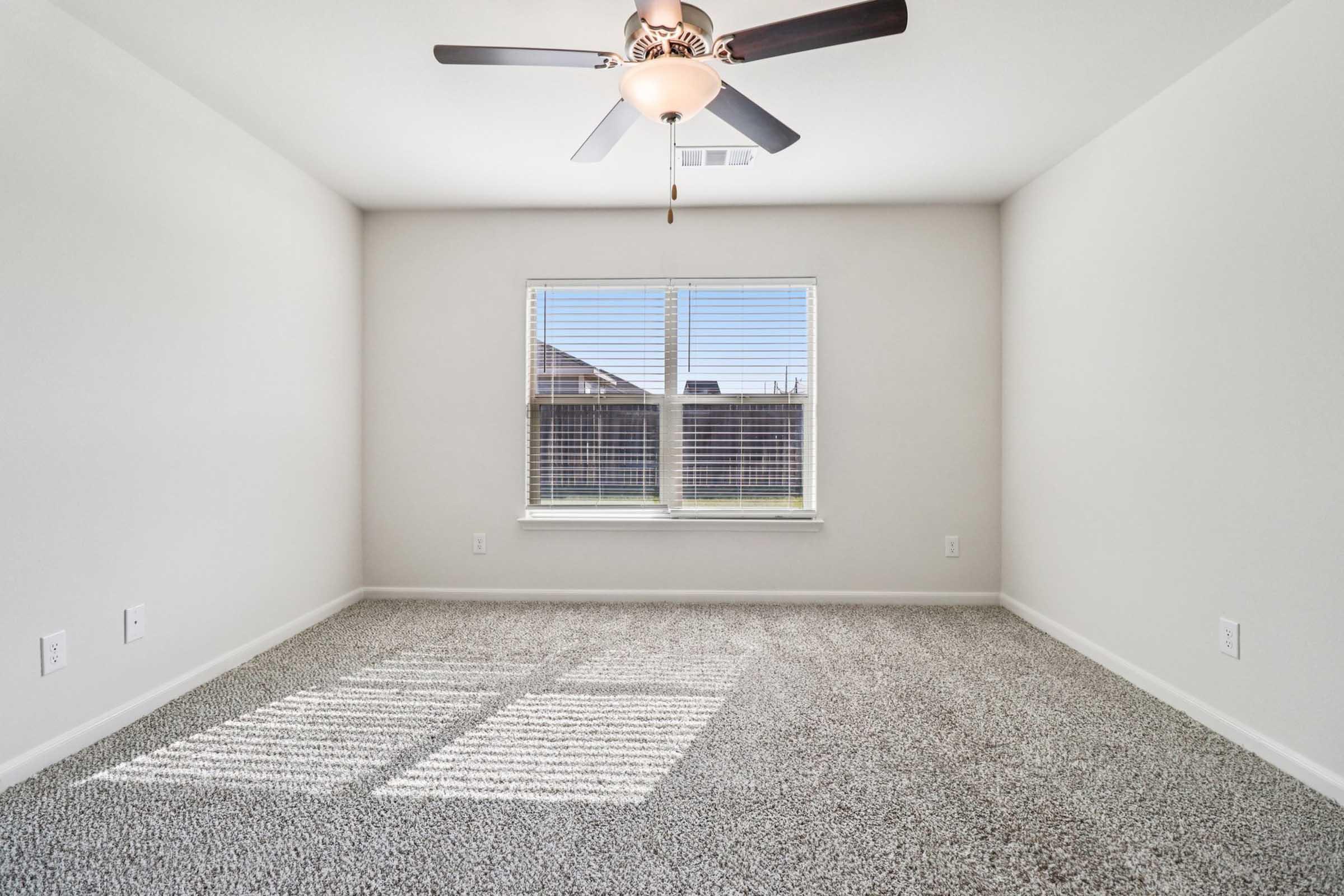 A well-lit, empty room with light gray walls and a ceiling fan. The floor is covered with soft, beige carpet. Two large windows let in natural light, showcasing a view of the outdoors, while the space appears clean and ready for furnishings.