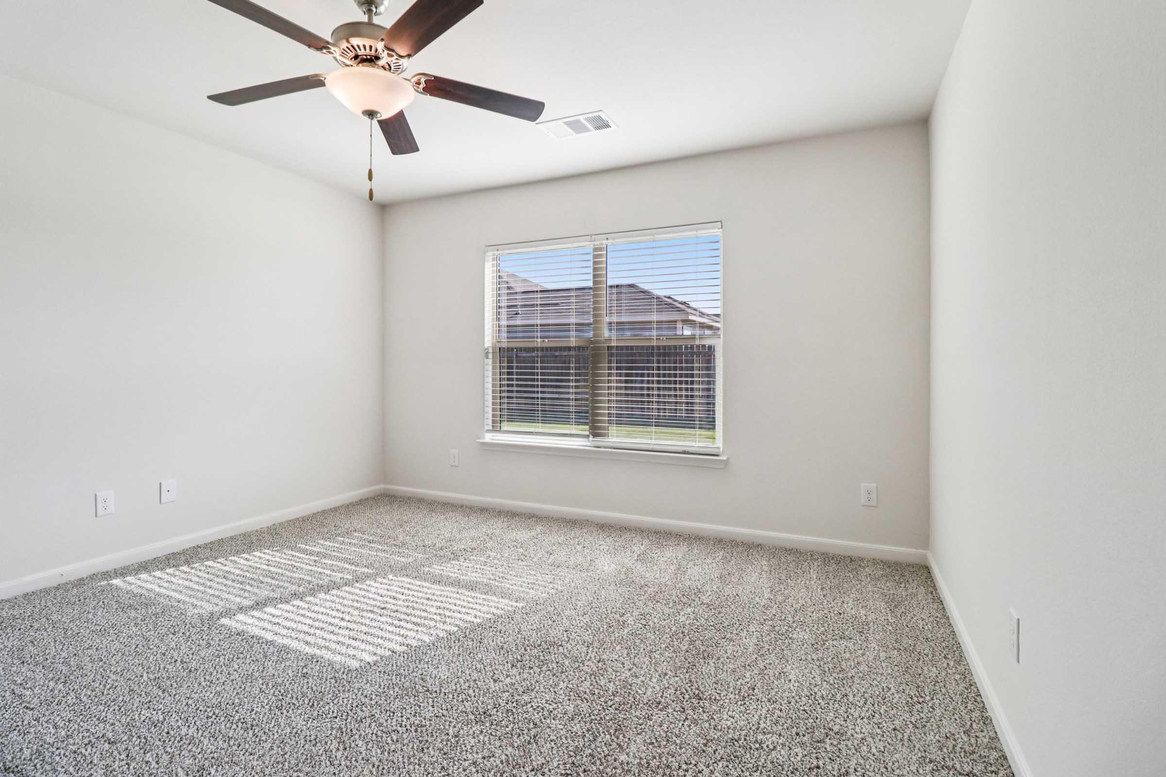 A well-lit empty room featuring light-colored walls, a ceiling fan with a light fixture, and a large window with blinds. The floor is covered in plush, light-colored carpet, and natural light casts shadows across the floor.