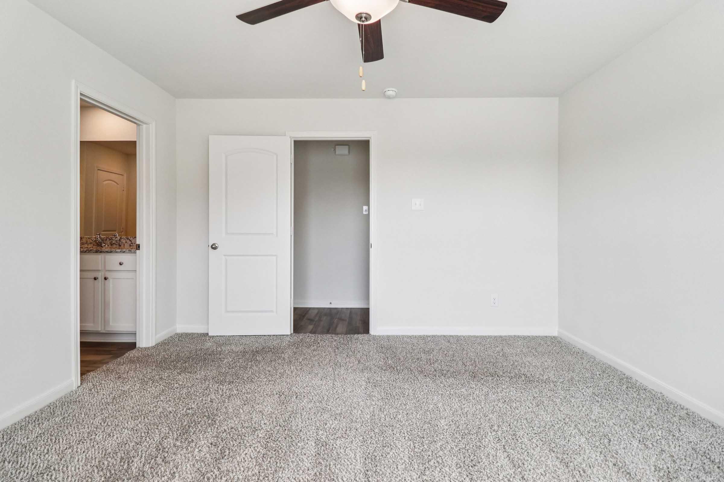 A bright, empty room with light-colored walls and carpets. There is a ceiling fan with wooden blades. On one side, a doorway leads to another space, possibly a bathroom. The room has a clean, modern feel with ample natural light.