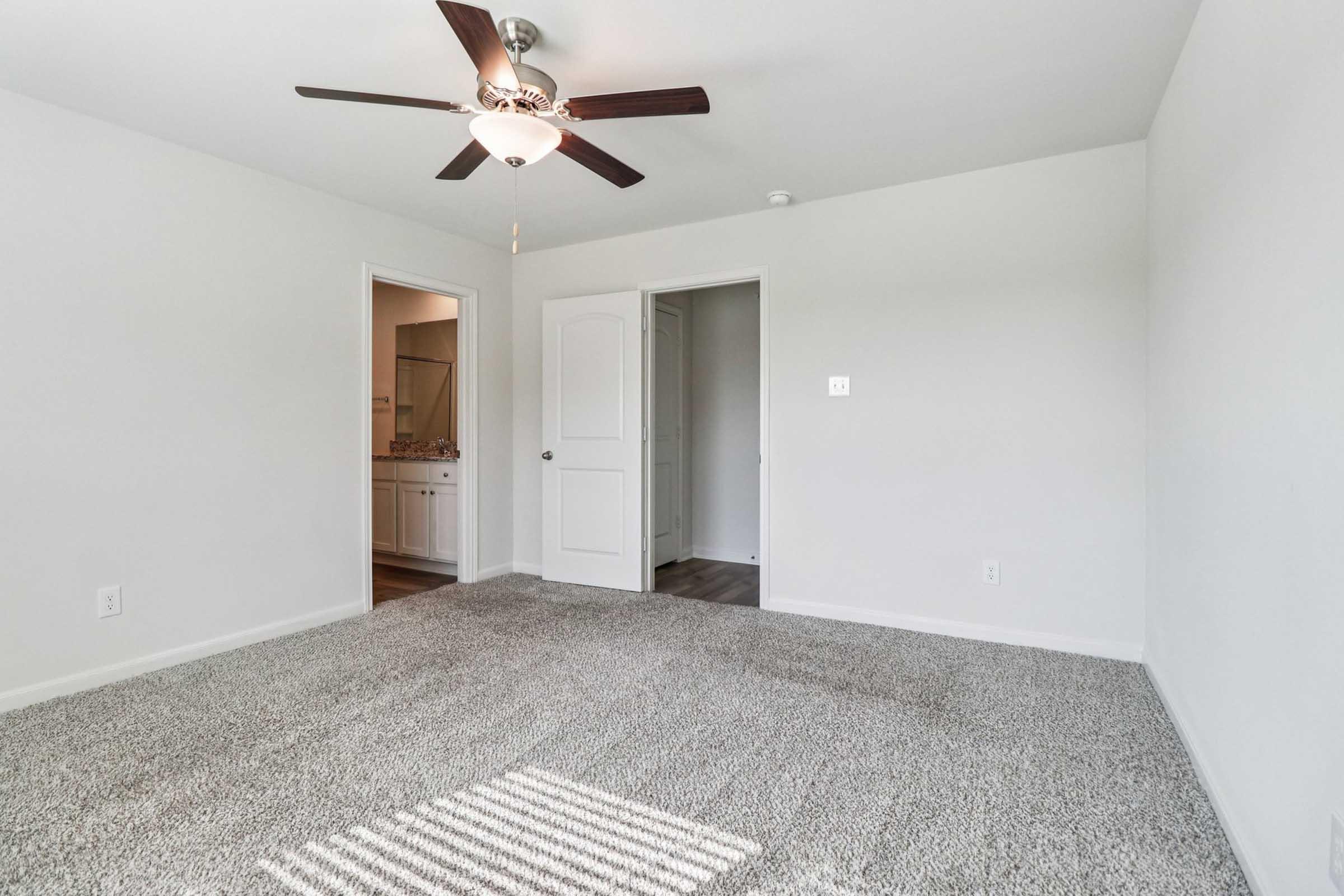 A spacious, well-lit room featuring light gray walls and plush carpeting. A ceiling fan is mounted above, and there are two doors leading to other spaces. Soft natural light filters through a window, creating a welcoming atmosphere. Ideal for a bedroom or cozy sitting area.