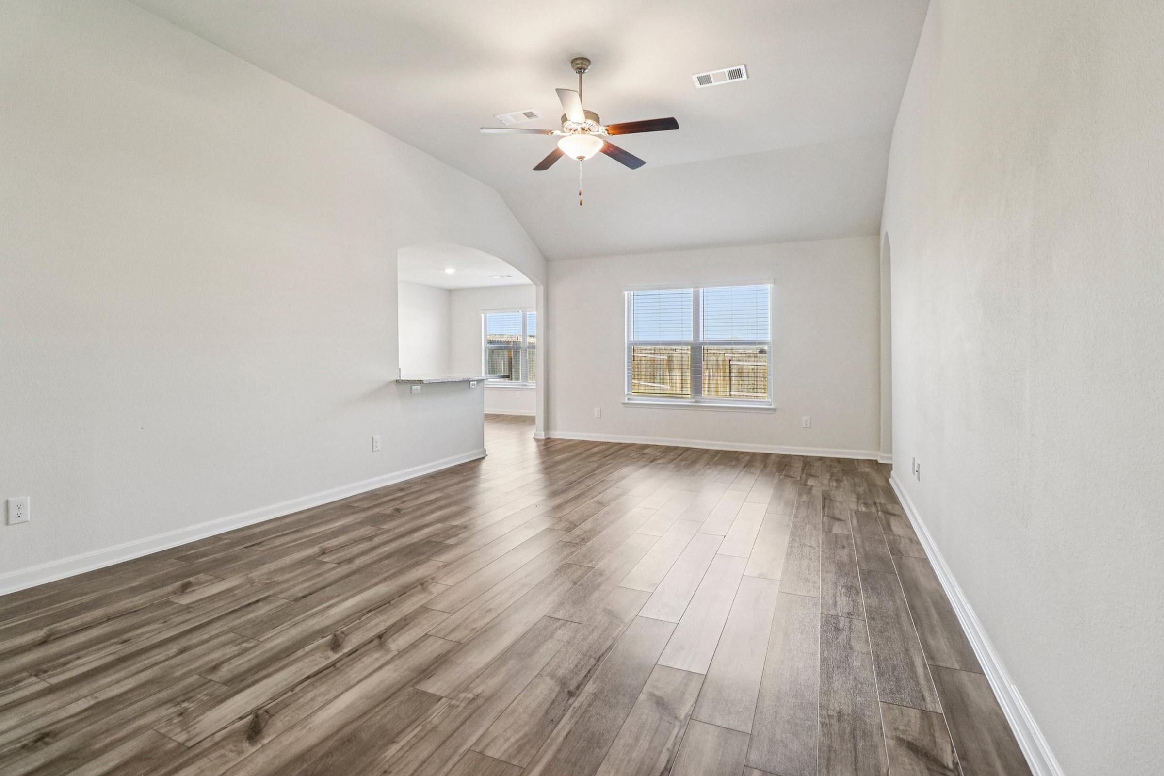 A spacious, well-lit living room with light wooden flooring and a ceiling fan. Large windows allow natural light to fill the room, and a small arch leads to a kitchen area in the background. The walls are painted a neutral color, creating a bright and inviting atmosphere.