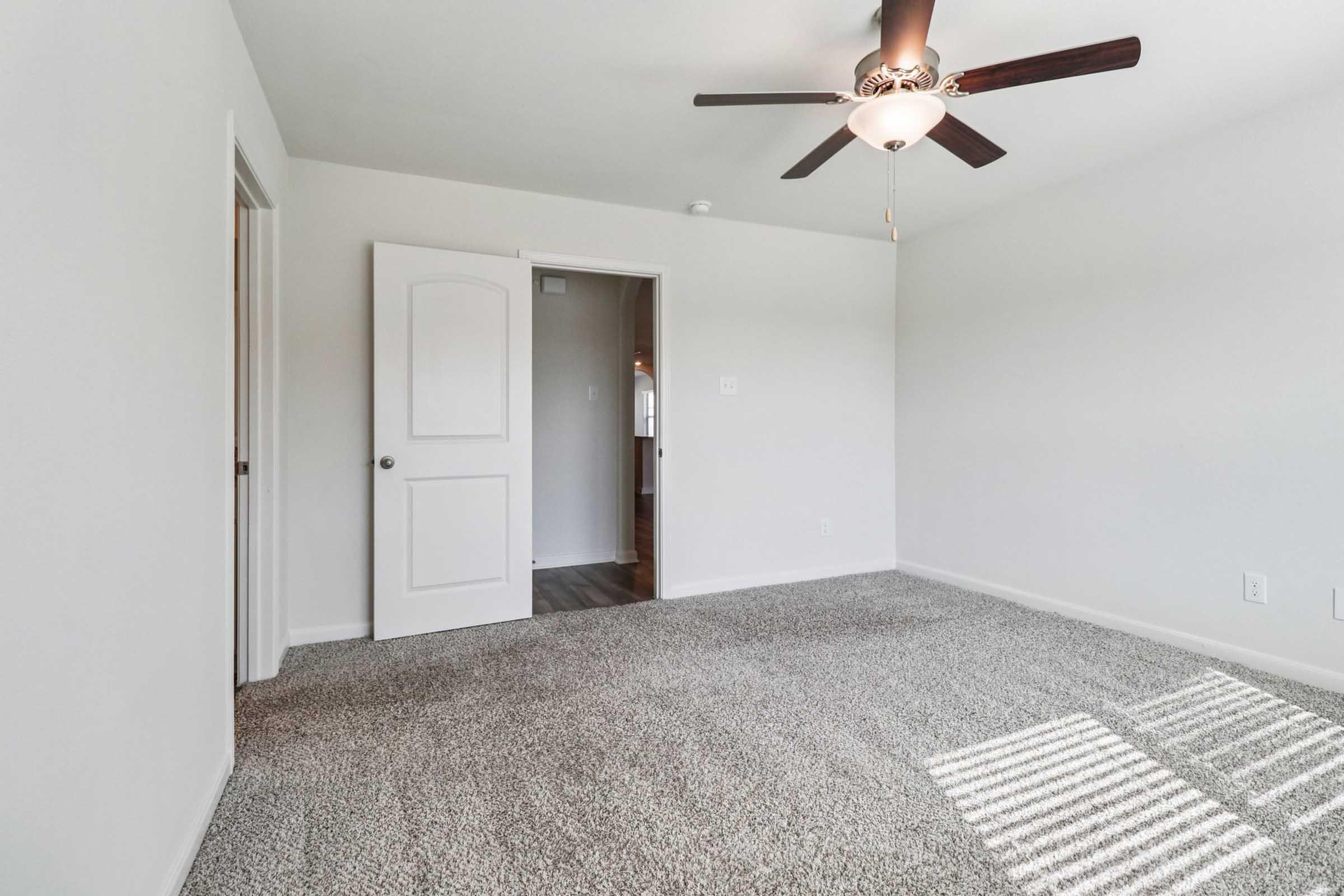 A spacious, well-lit room featuring light gray walls and a plush carpet. A ceiling fan is mounted above, and there's a closed door on the left and an open doorway leading to another room in the background. Sunlight casts soft shadows on the floor, creating a warm and inviting atmosphere.