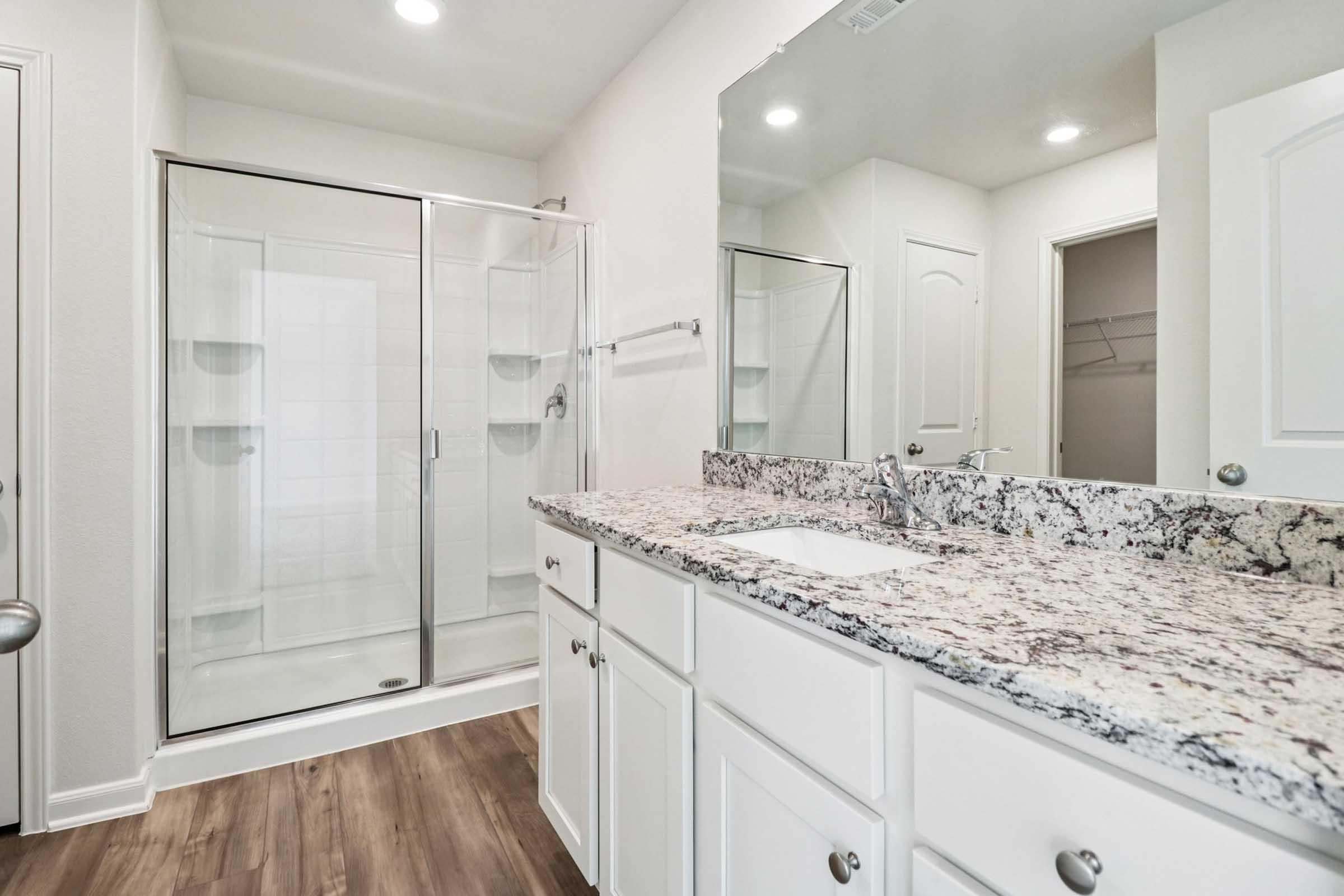 A modern bathroom featuring a glass-door shower, white cabinetry with a granite countertop, and wooden flooring. The room is well-lit with recessed lighting and a large mirror above the sink. A door on the left leads to another space, and the walls are painted a neutral color.
