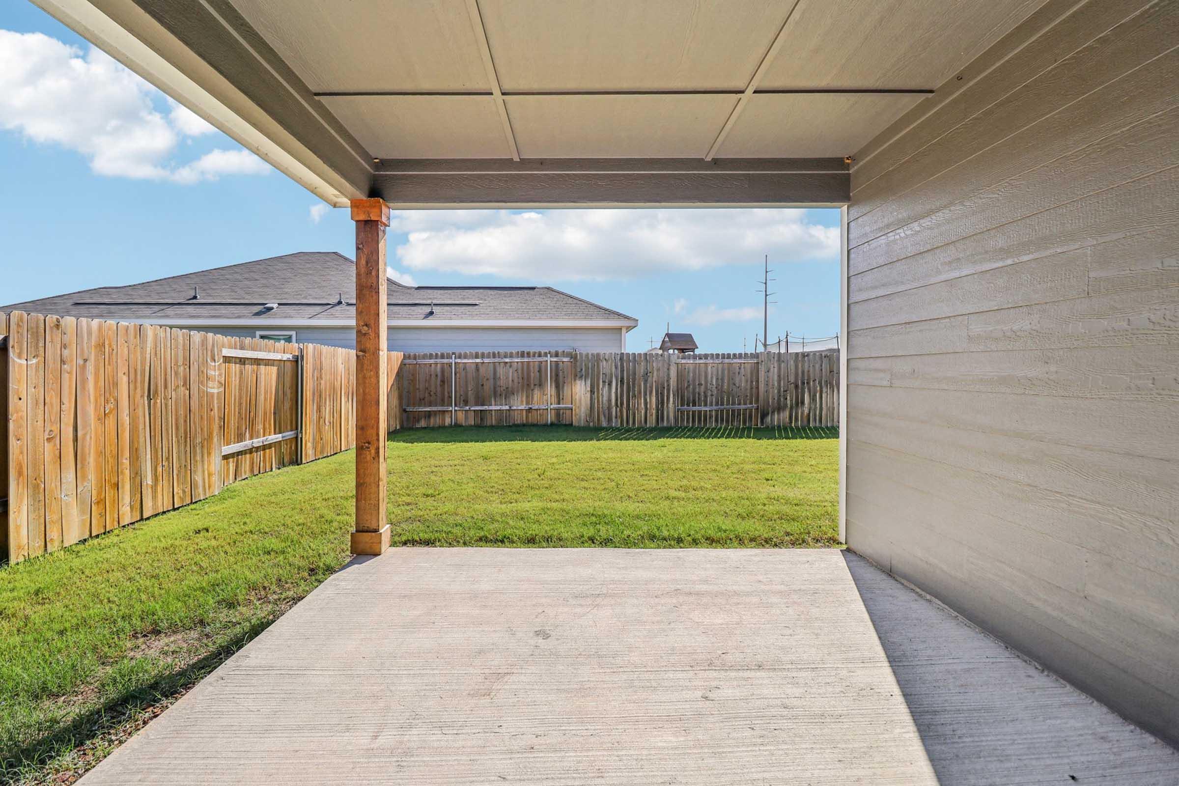 View from a covered patio looking out onto a grassy backyard enclosed by a wooden fence. The sky is clear with a few clouds, and a neighboring house is visible in the background. The patio has a concrete floor.
