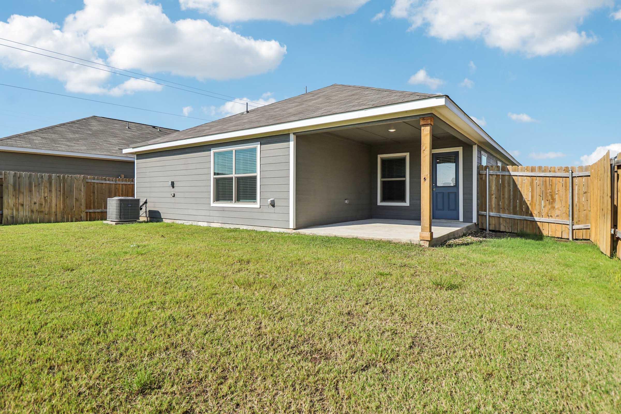 A single-story house with a grey exterior and a covered patio. The backyard features green grass and a wooden fence, while an air conditioning unit is visible on the side. The sky is partly cloudy, creating a bright atmosphere.