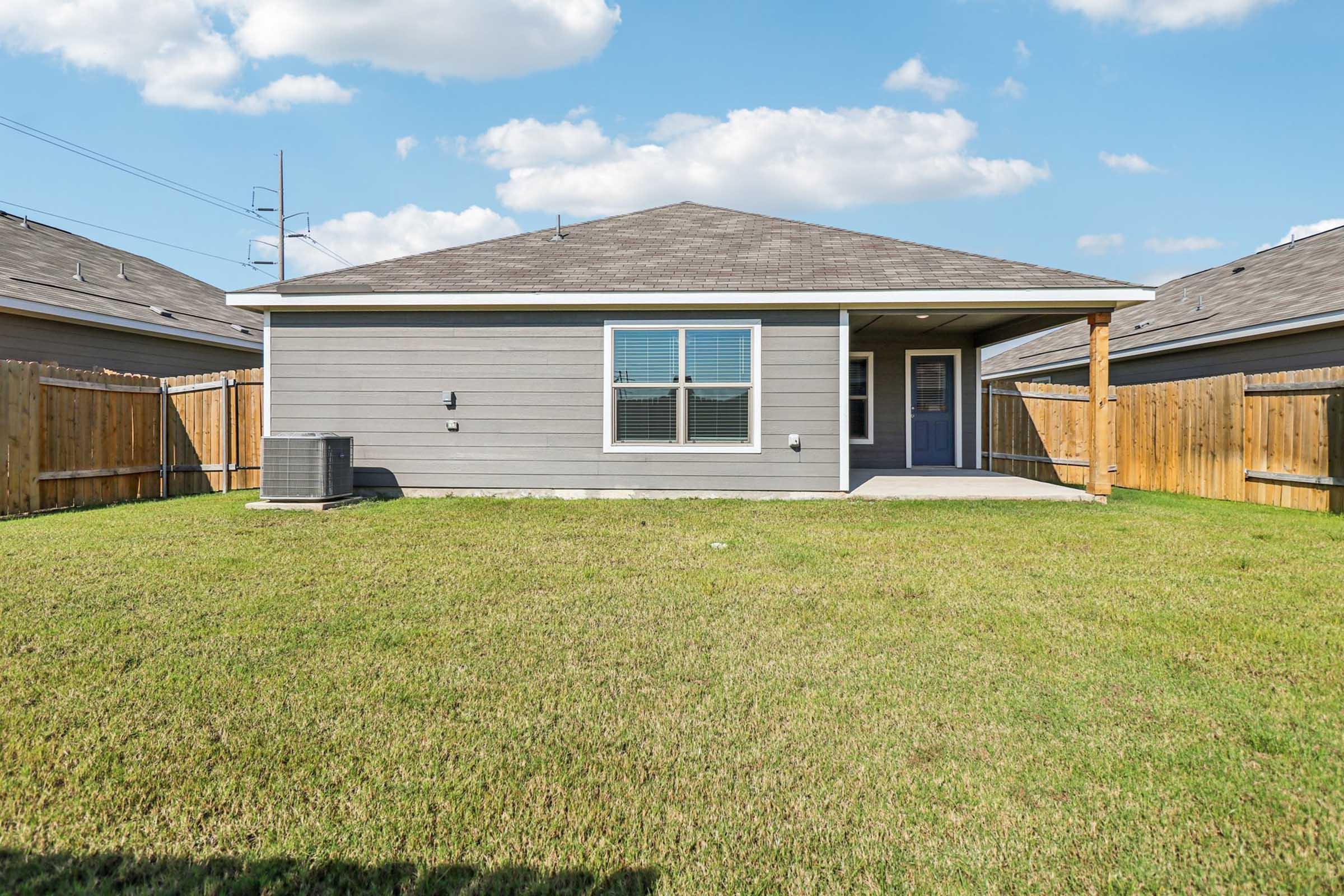 A single-story house with a gray exterior and blue door, set in a spacious backyard. The yard is well maintained with green grass, and there is an air conditioning unit on the side of the house. A wooden fence surrounds the backyard, and the sky is partly cloudy.