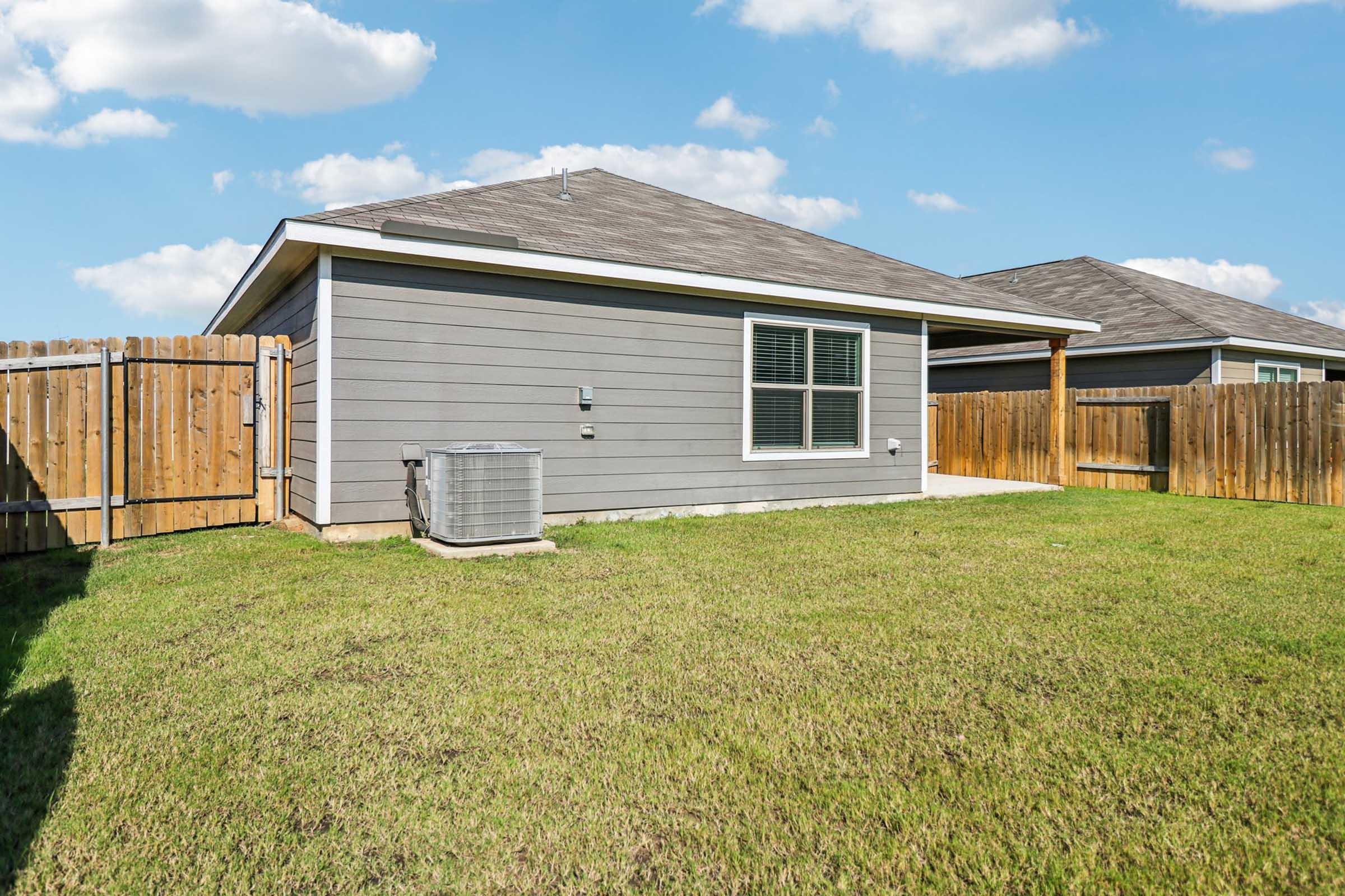 A view of a backyard featuring a single-story gray house with a sloped roof, an air conditioning unit on the side, and a fenced yard with green grass under a clear blue sky. Nearby, another house is visible in the background behind the fence.
