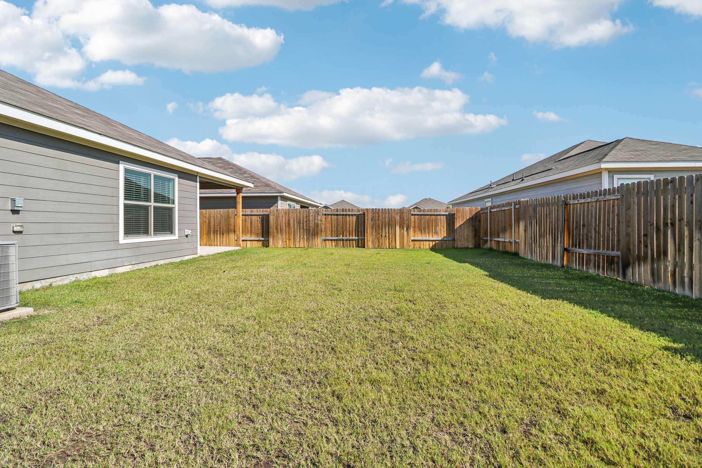 Well-maintained backyard showing a grassy area enclosed by wooden fences. The scene features two neighboring houses on either side, with blue skies and fluffy white clouds above.